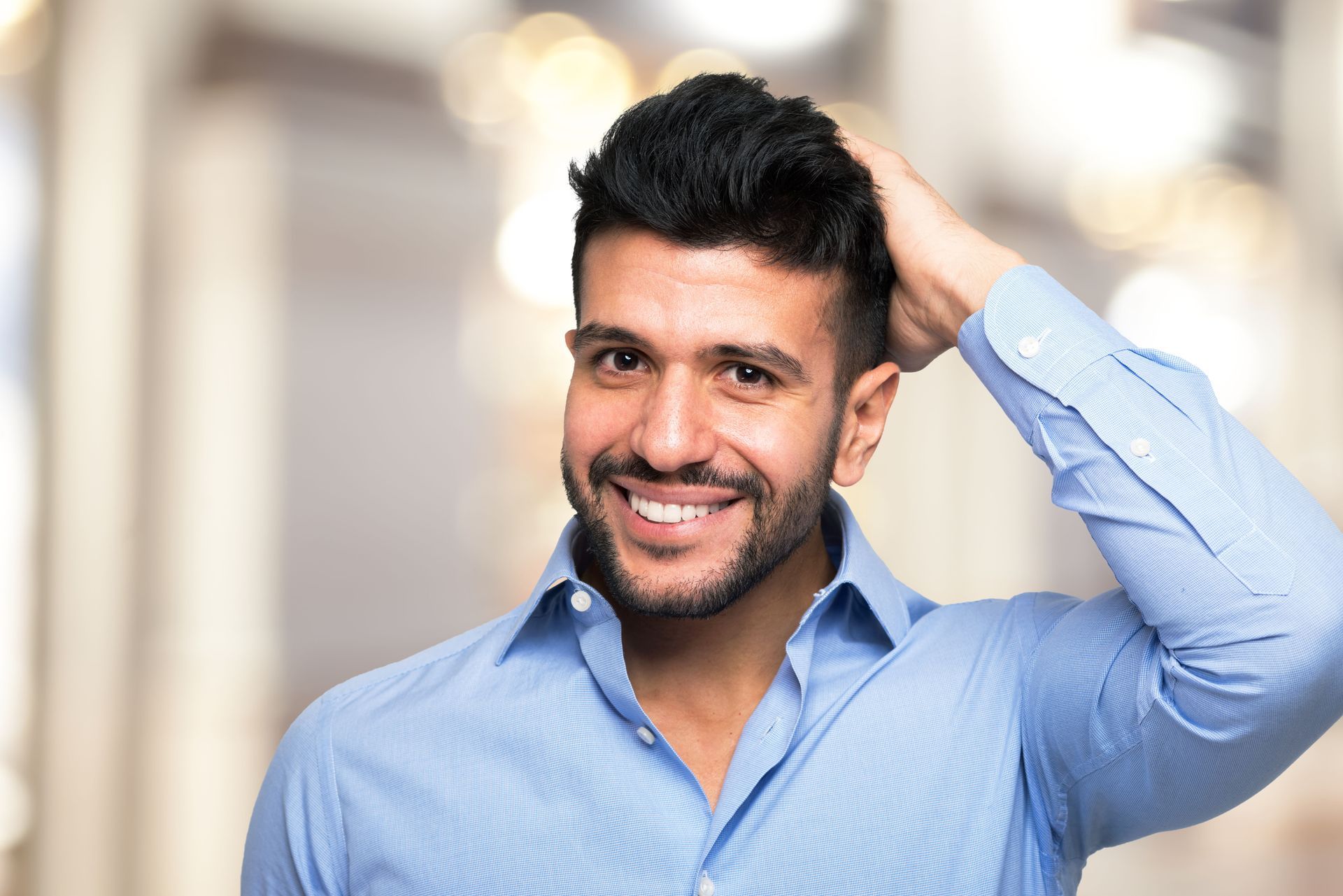 Man in blue shirt smiling, hand in hair, blurry background.