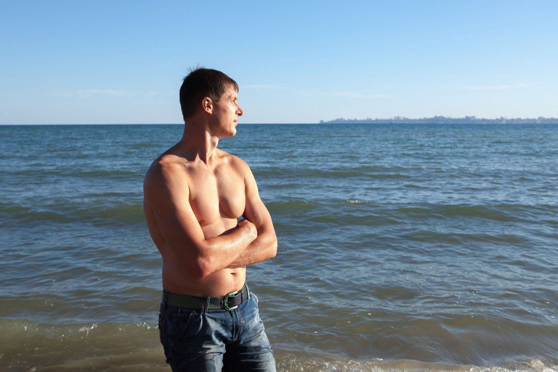 Man with bare chest standing at the beach, arms crossed, looking out at the water.