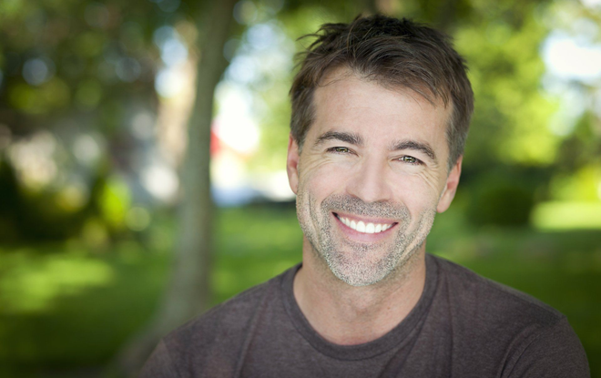 Man with a graying goatee smiles broadly outdoors; blurred green foliage in background.