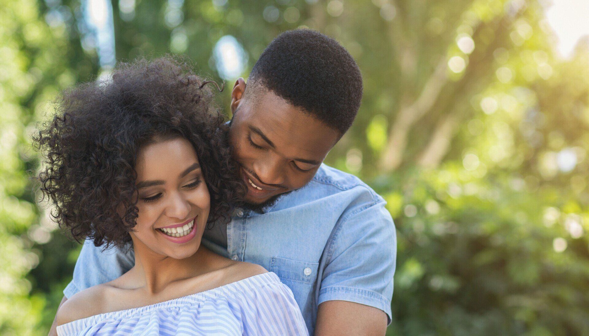 Man embracing a smiling woman outdoors; sunlight through trees.