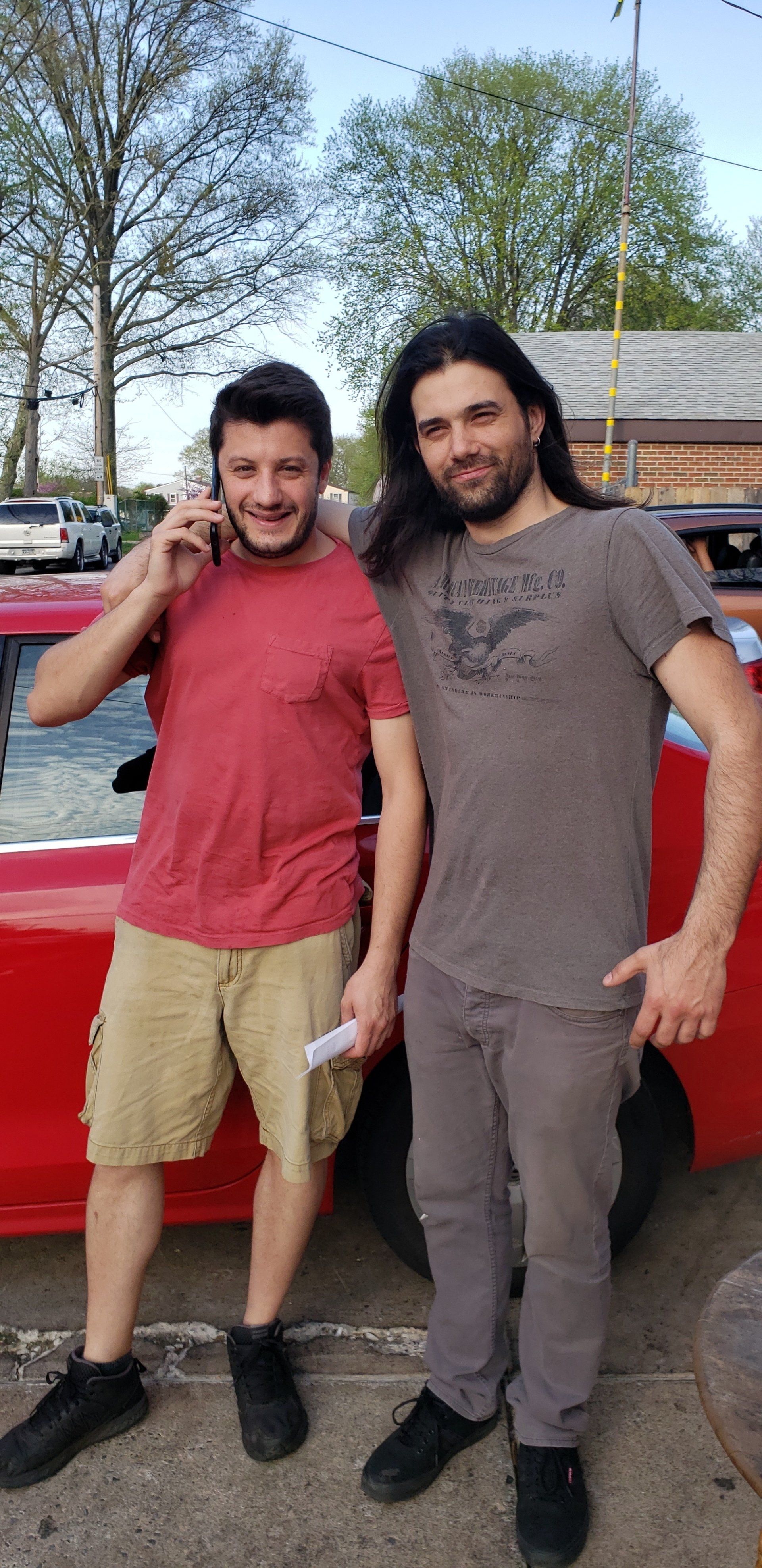 Two men are standing next to each other in front of a red car.