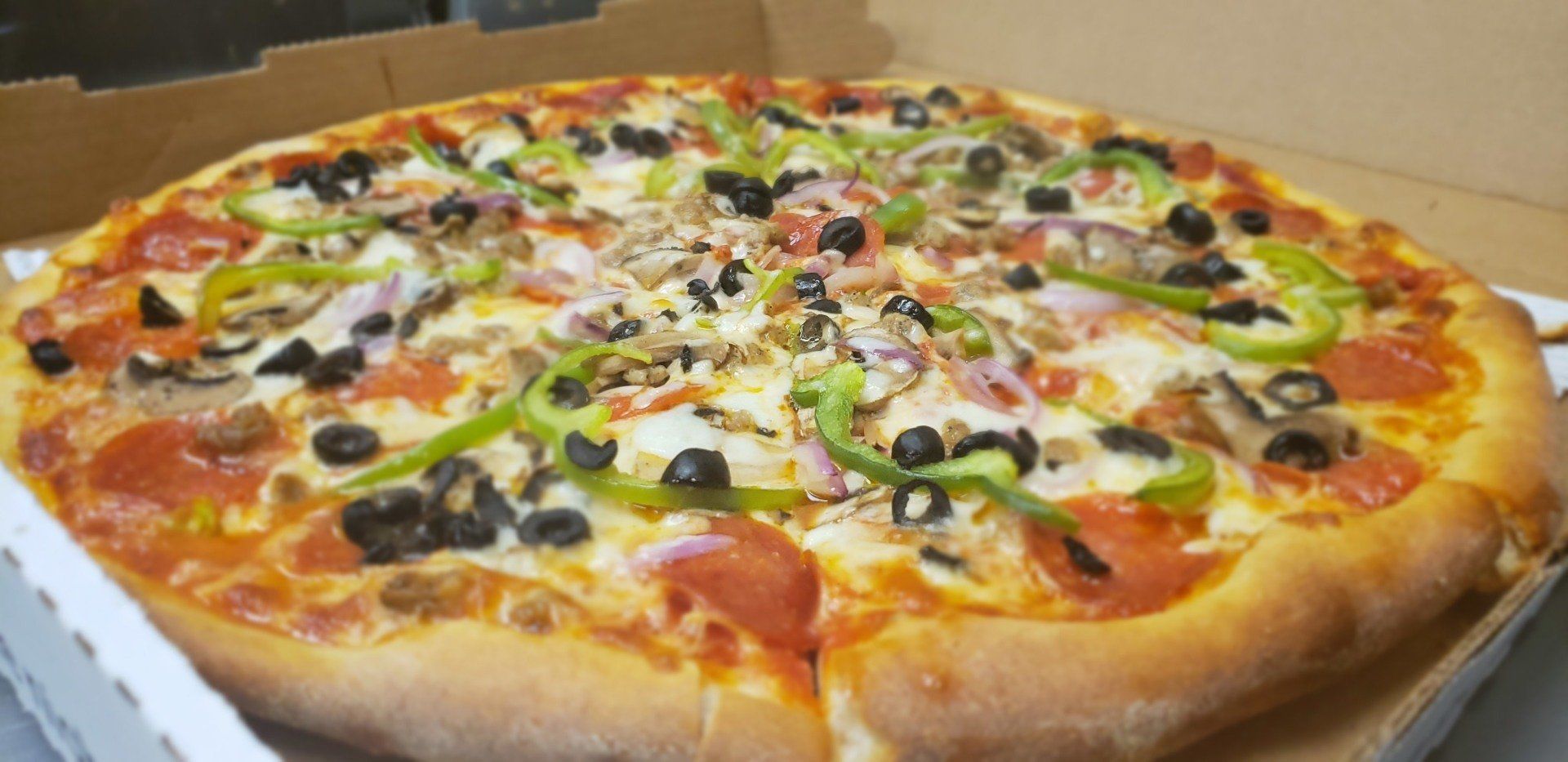 A pizza with pepperoni , olives , peppers , onions and cheese is in a box.