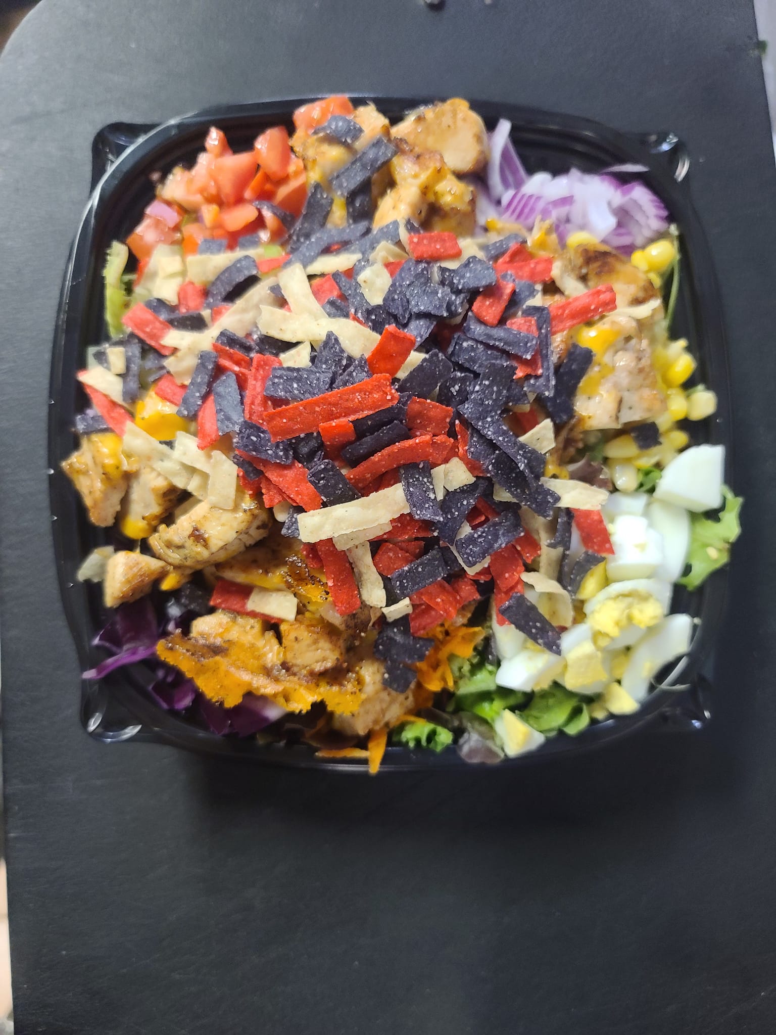 A salad with chicken , tortilla chips , eggs , and other vegetables in a black container.