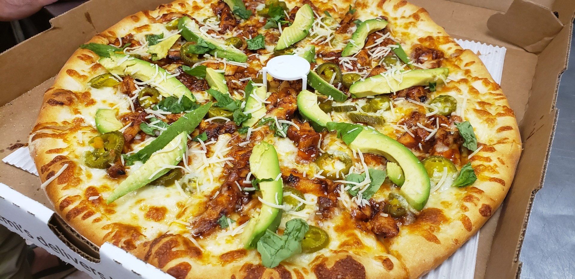 A pizza with avocado , jalapenos and cheese is in a box.