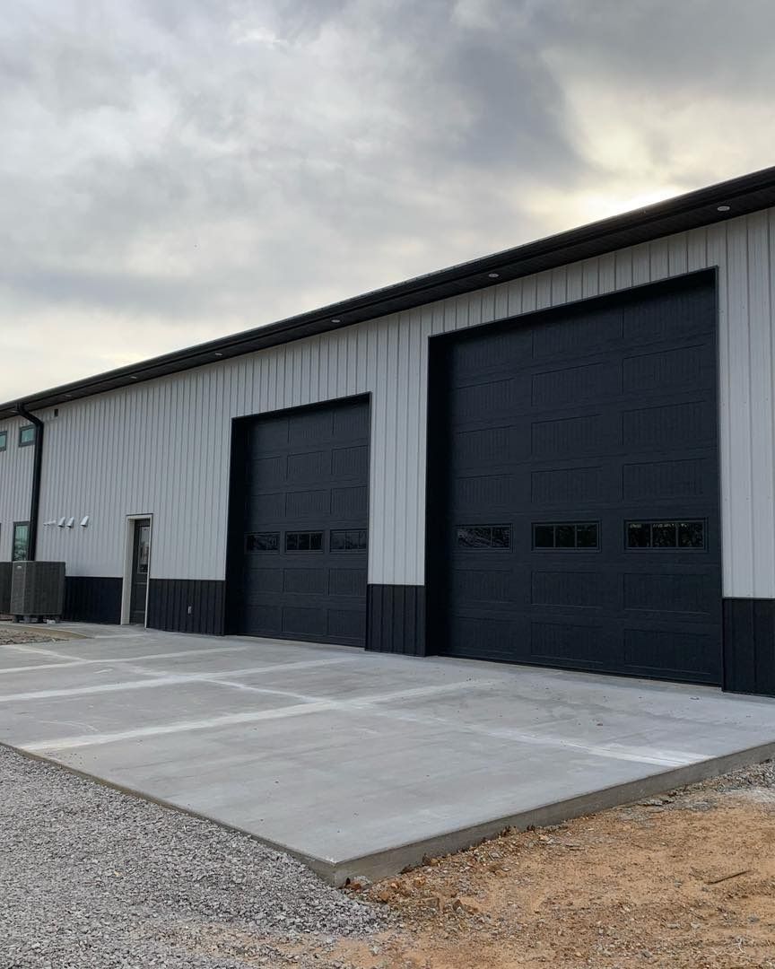 Exterior view of a metal building with two black garage doors and a concrete pad.