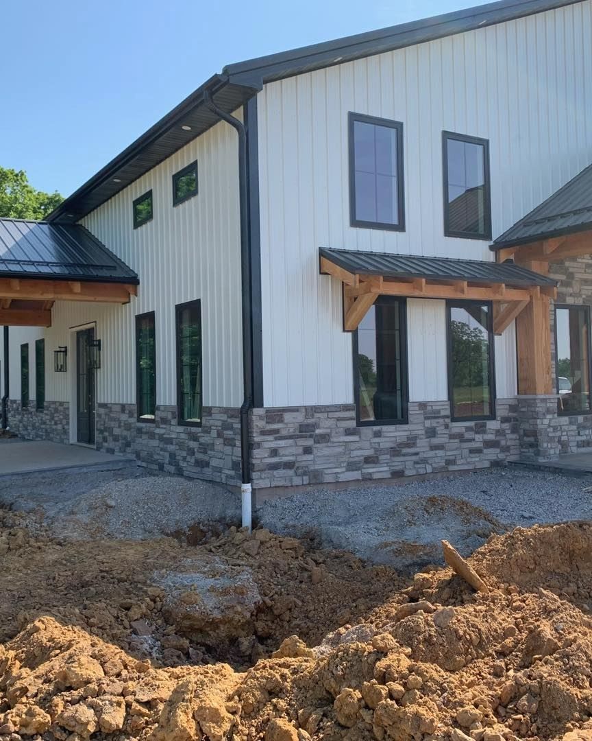 Modern white building with black trim, windows, and stone base. Dirt and gravel in the foreground.