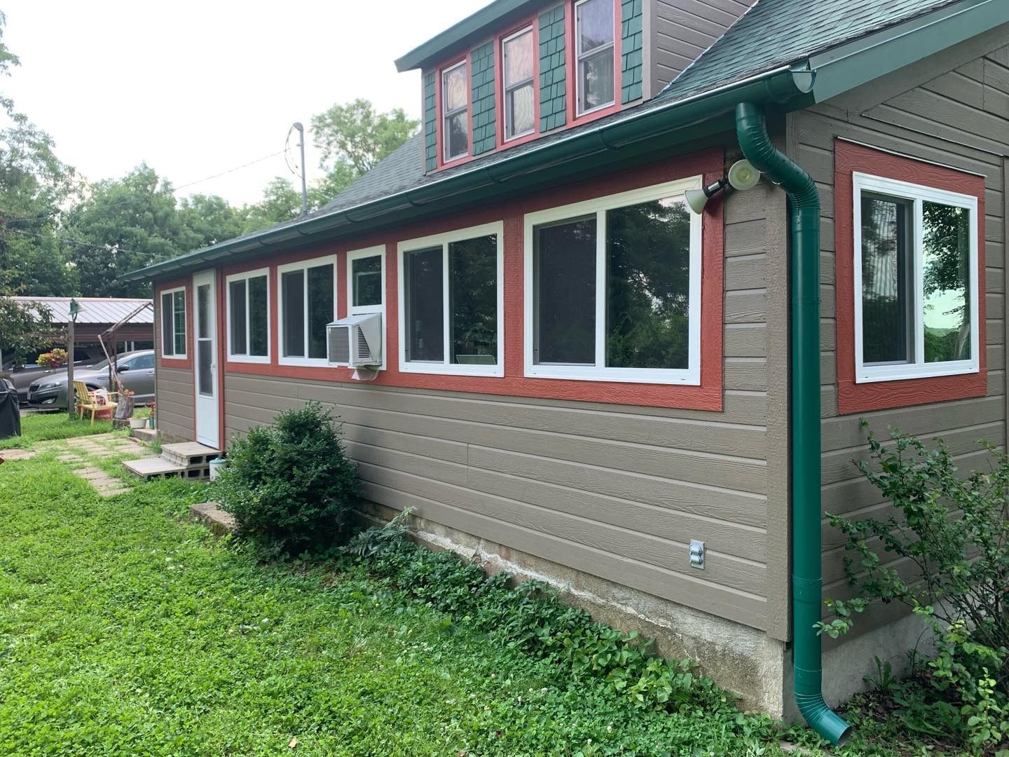 Side view of a house with green roof and trim, gray siding, and many windows. Green grass in foreground.