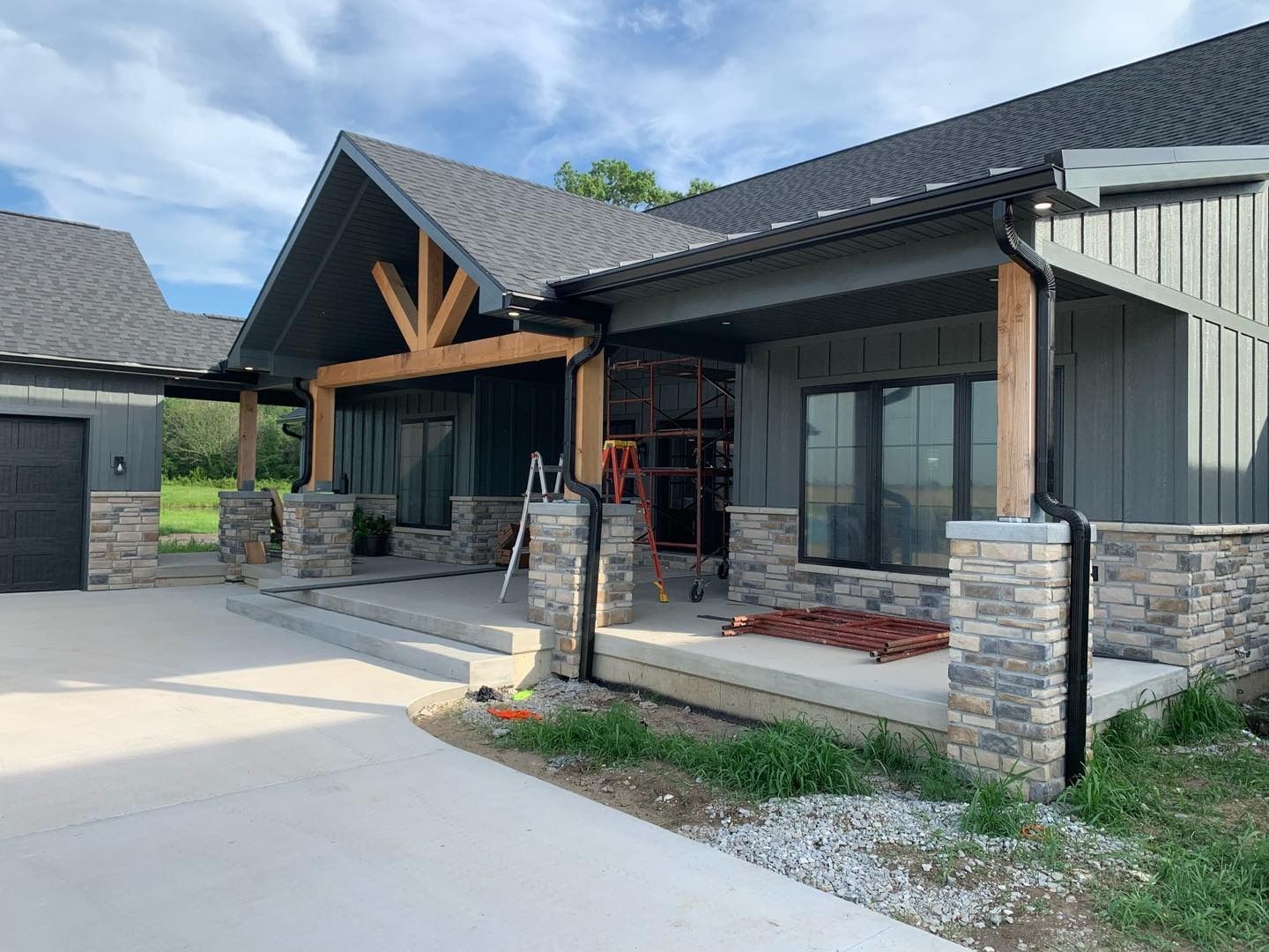 Gray and stone exterior of a modern house under construction. Porch with wood beams, windows, and gray siding.