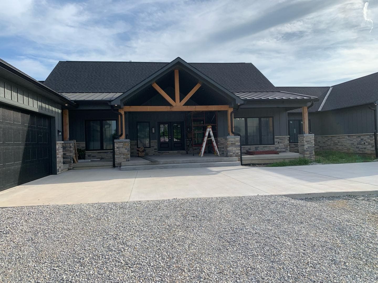 Dark gray house with wood accents, front porch, and gravel driveway under a cloudy sky.