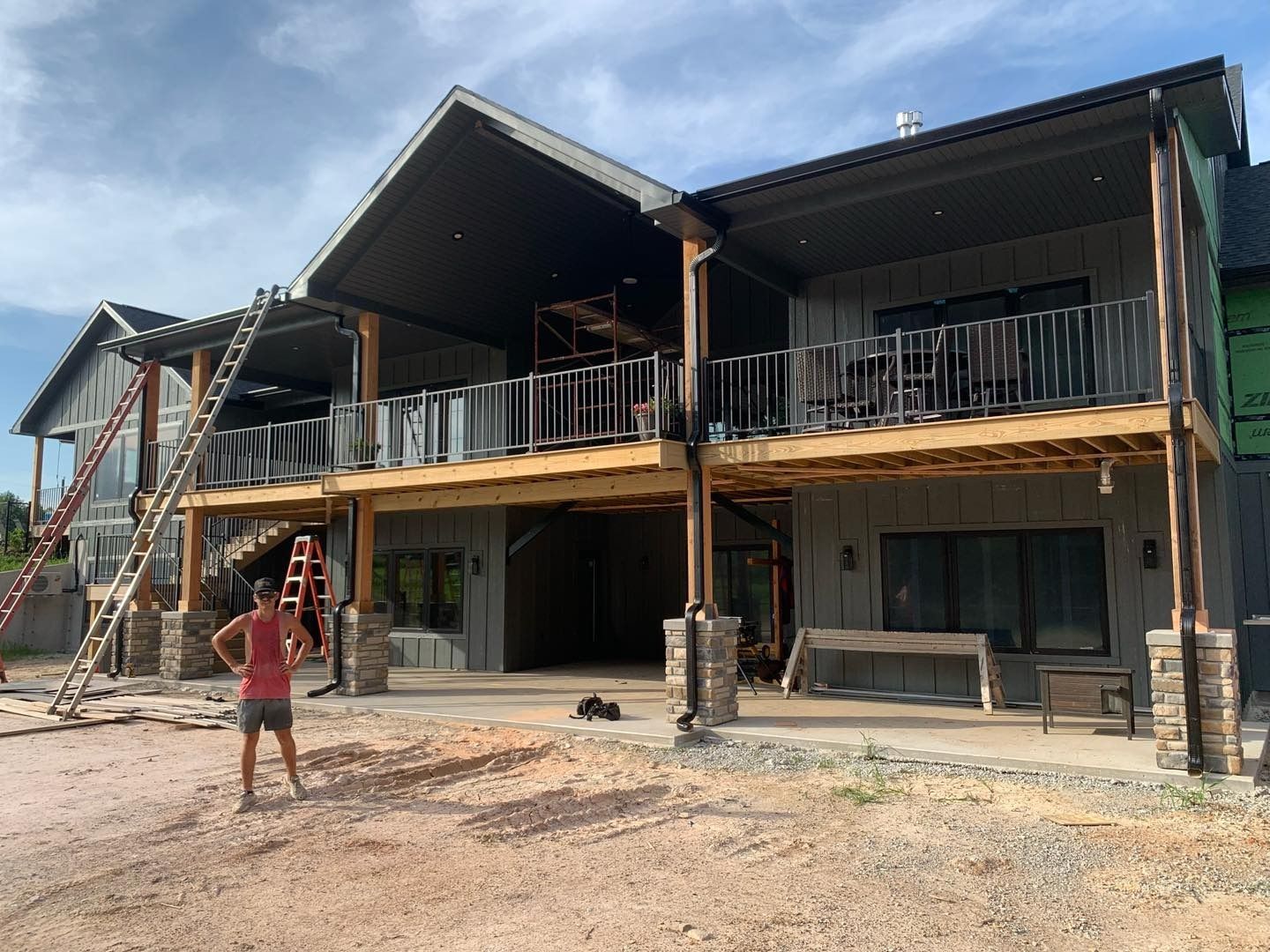 Two-story house under construction with a person standing in front. Gray siding, balconies, exposed wood posts, and a ladder are visible.