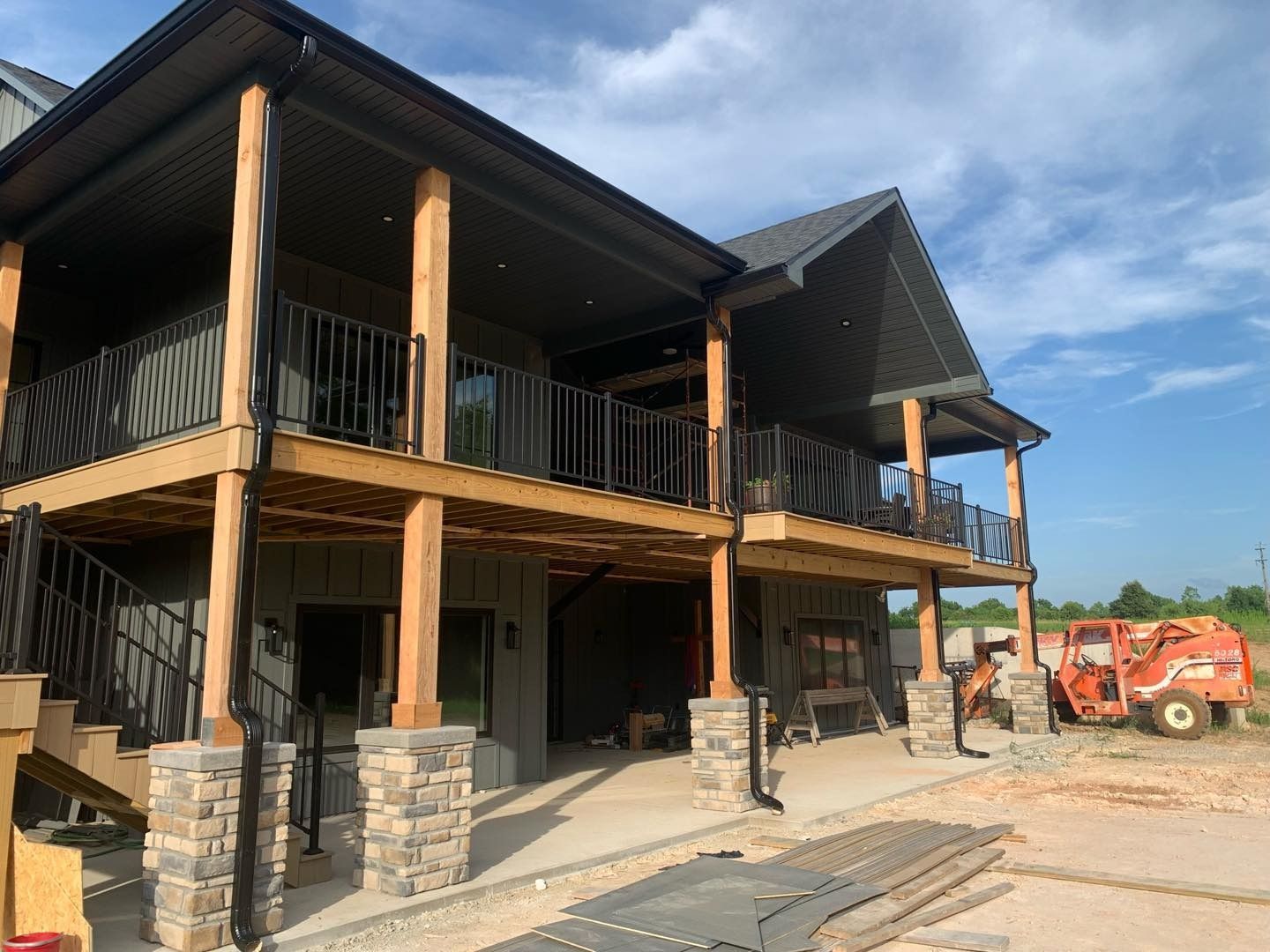 Two-story house with wooden beams, black railings, and stone columns. Construction equipment visible.