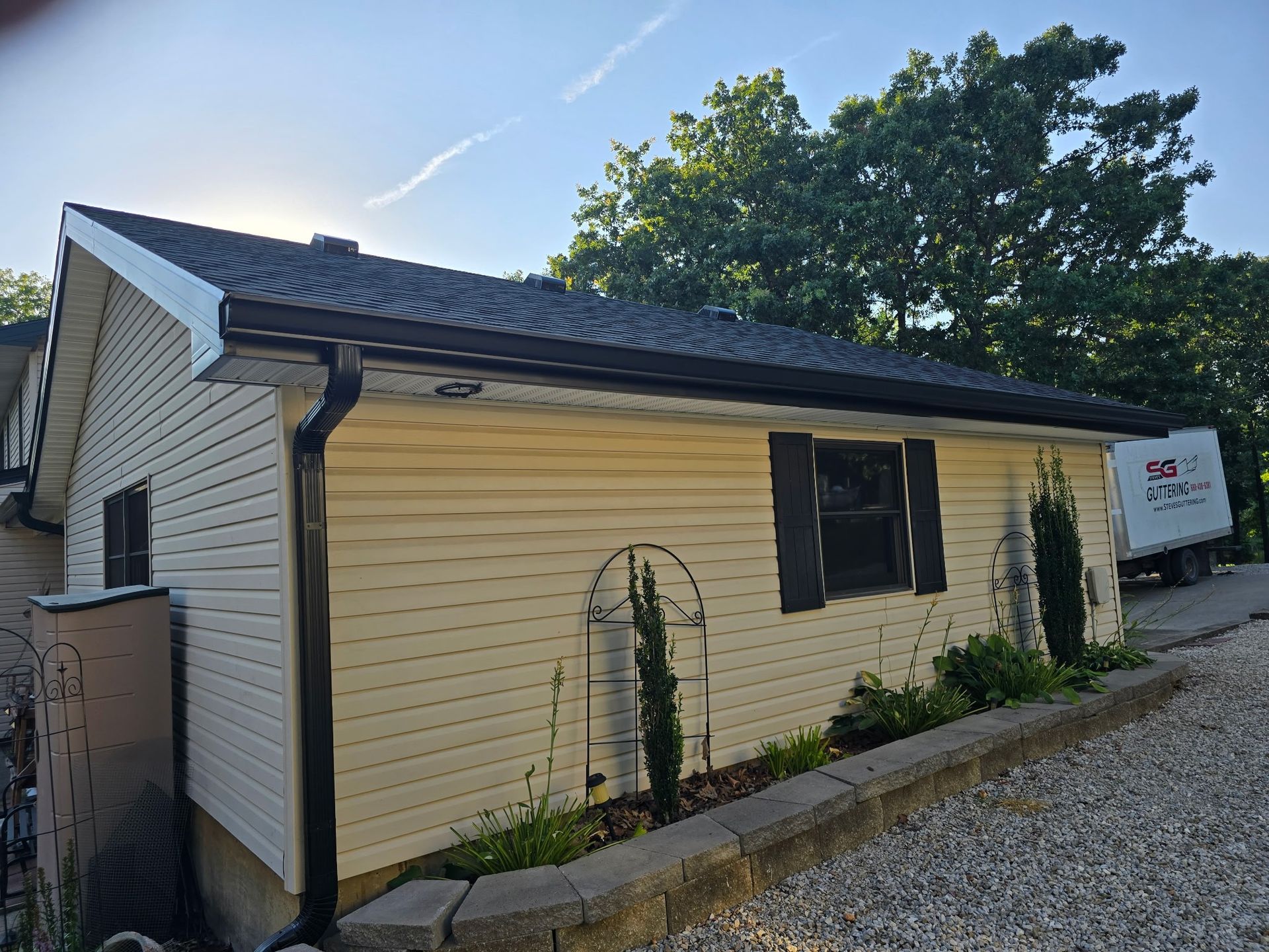 Tan sided building with black roof, gutters and window shutters, small garden, gravel driveway.