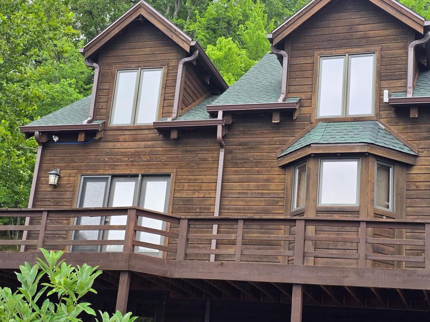 Two-story wooden house with deck and green roof against a backdrop of trees.