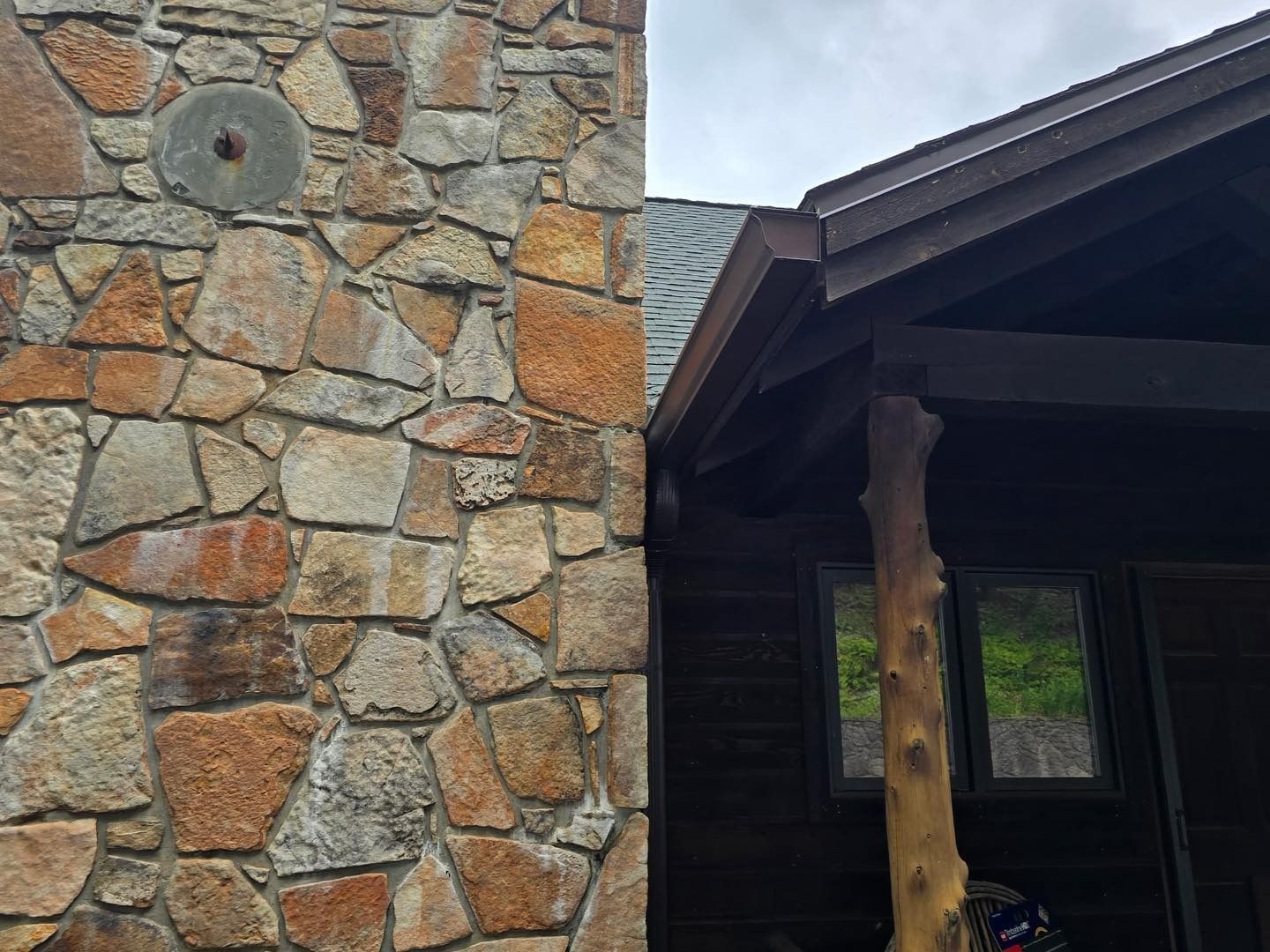 Stone chimney next to wooden cabin with green roof, tree trunk support.