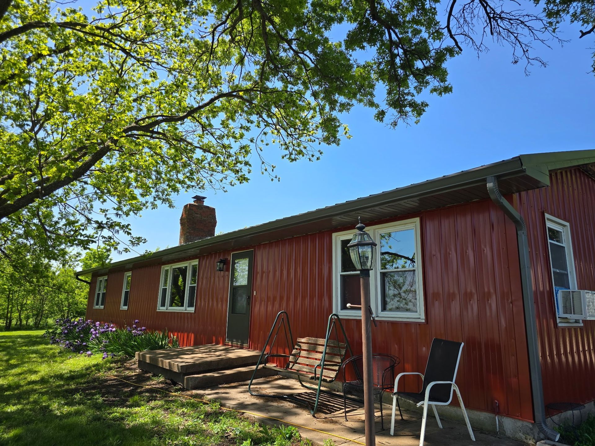 Red-sided house with a dark door, stone steps, and windows. Green trees and blue sky.