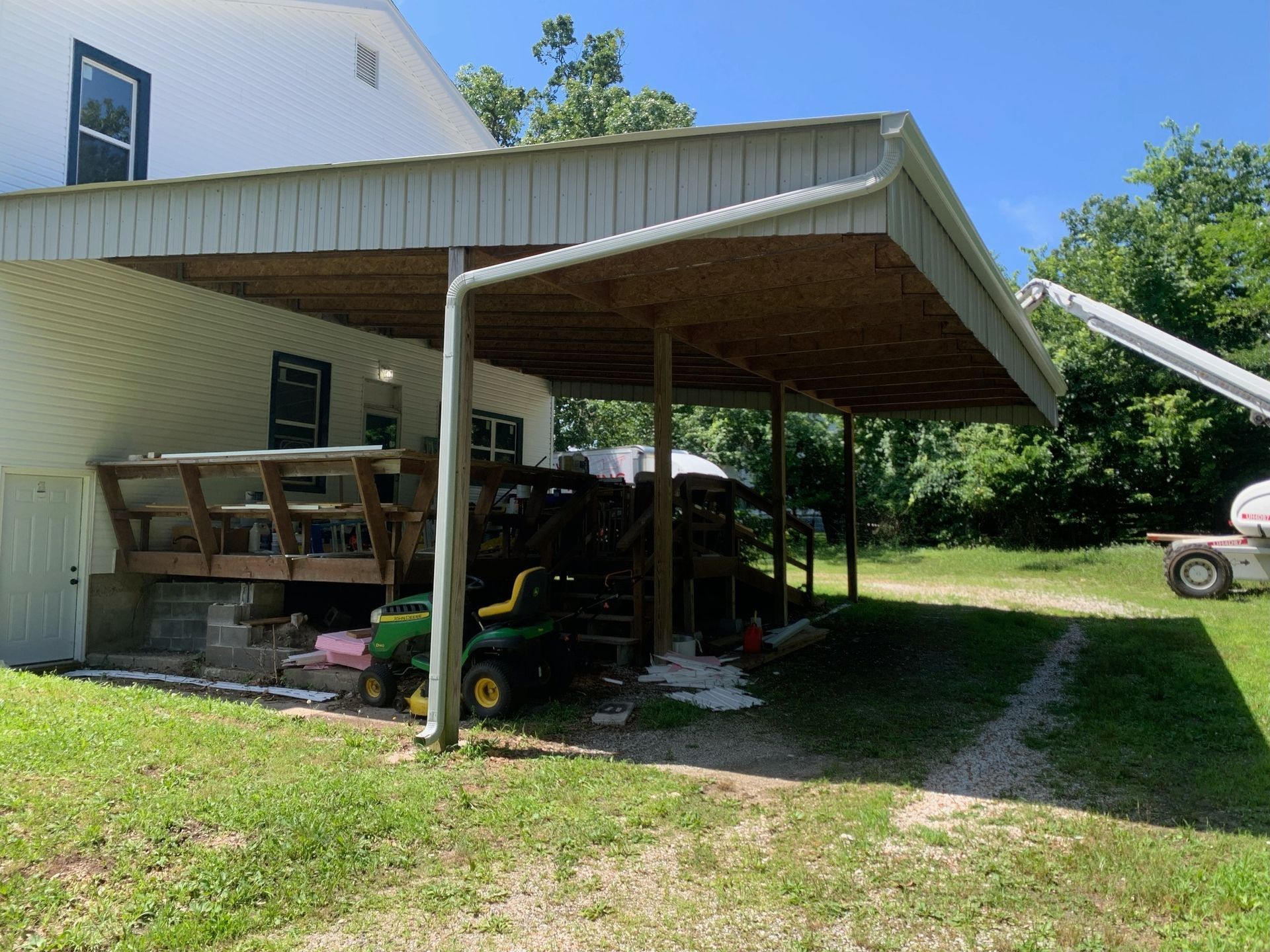 Carport attached to a house; lawn, deck, and tractor visible. Overcast roof supported by poles. Sunny, outdoor setting.