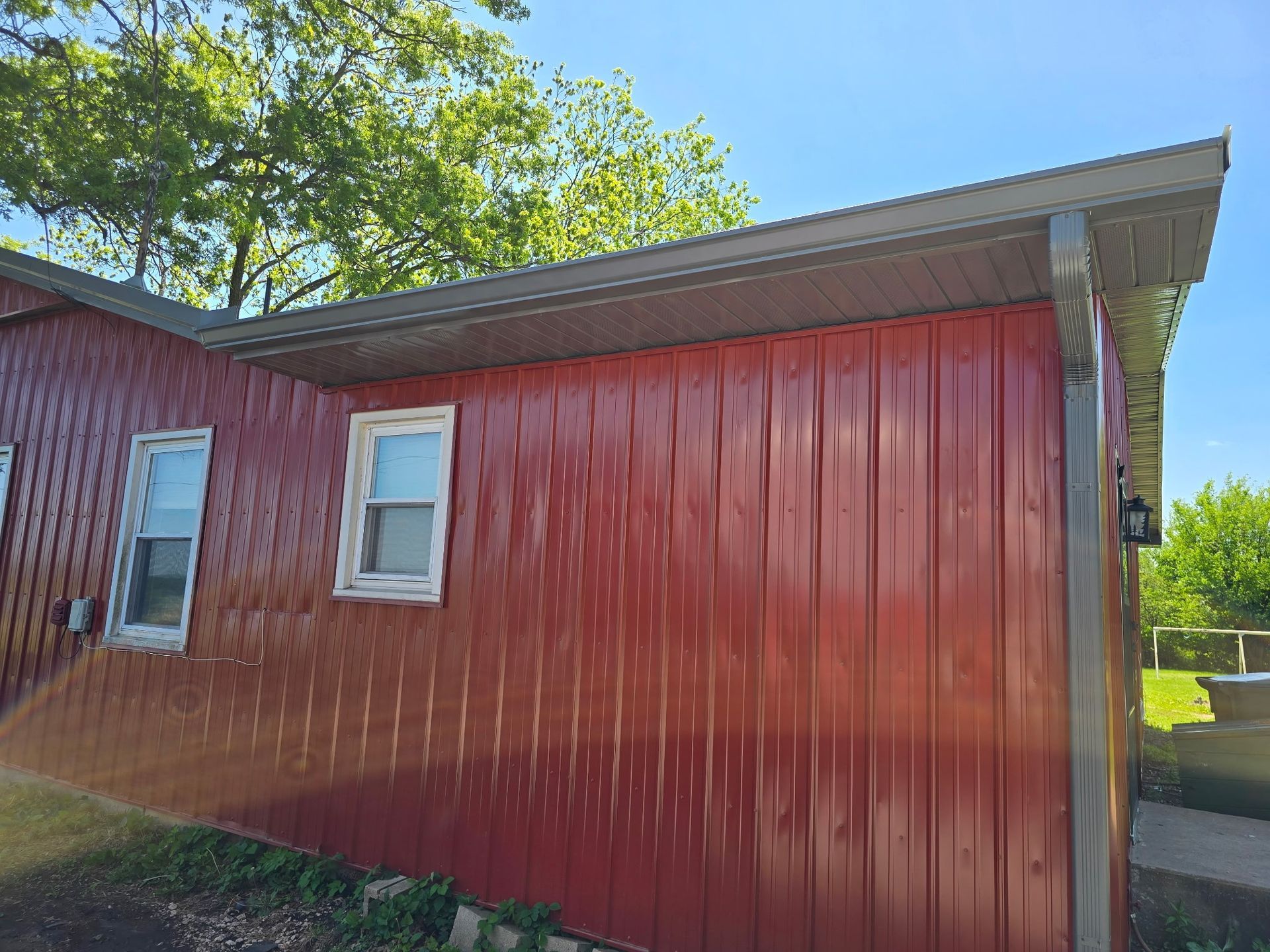 Red-sided building with two windows, a gray roof, and green foliage against a blue sky.