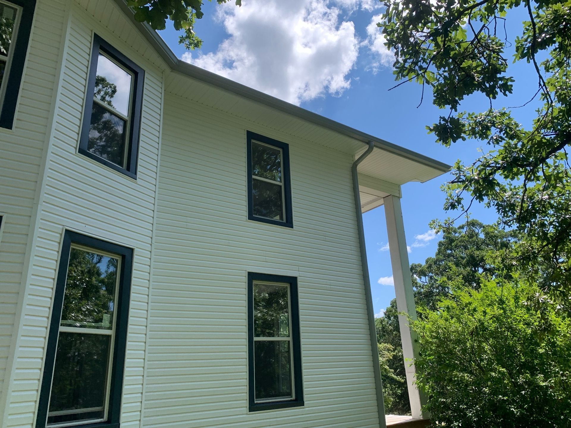 Side of a white house with dark blue window frames, surrounded by green trees under a blue sky with clouds.