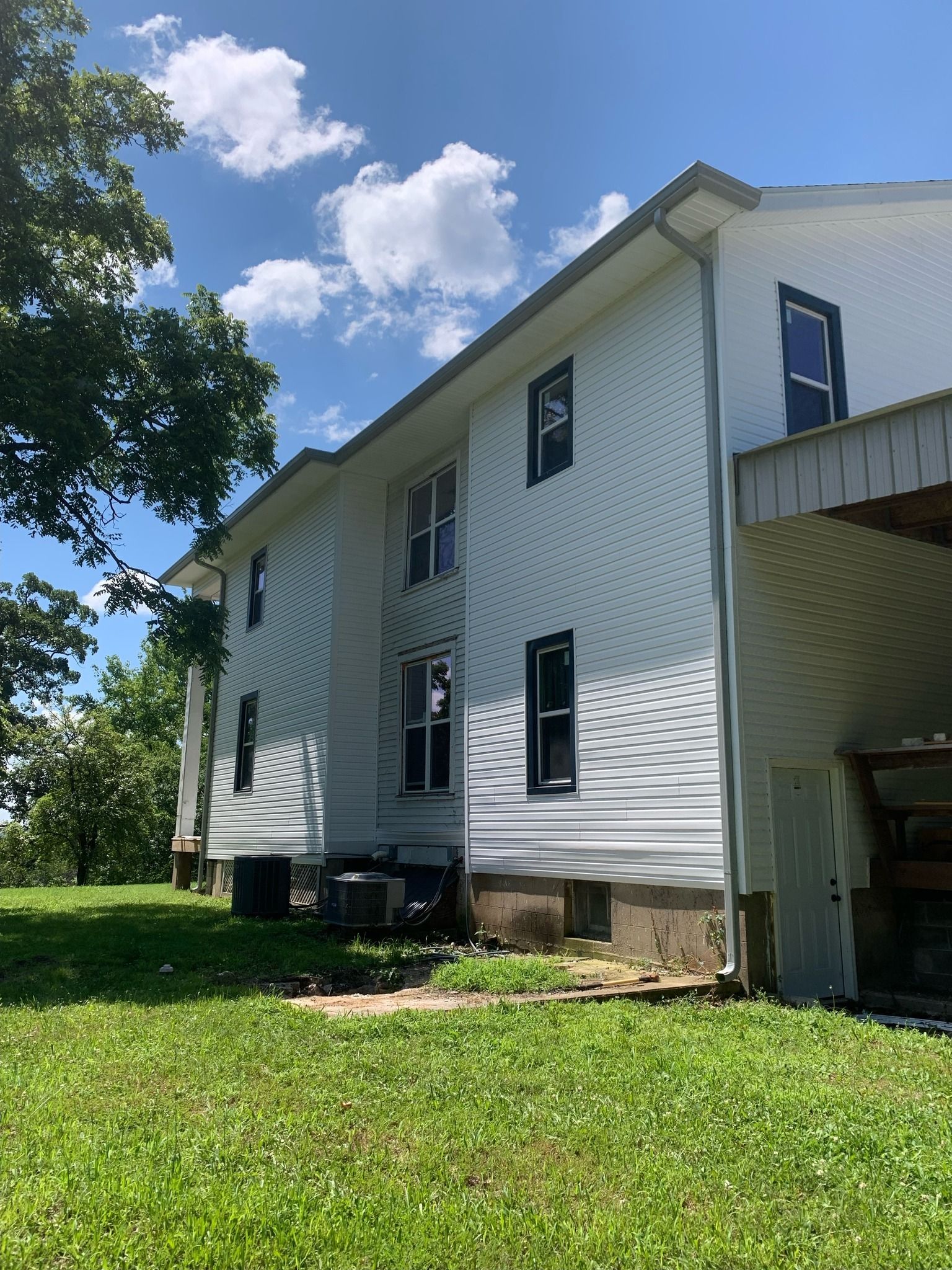 White two-story house with black window frames, side view, on a grassy lawn with trees and a cloudy blue sky.