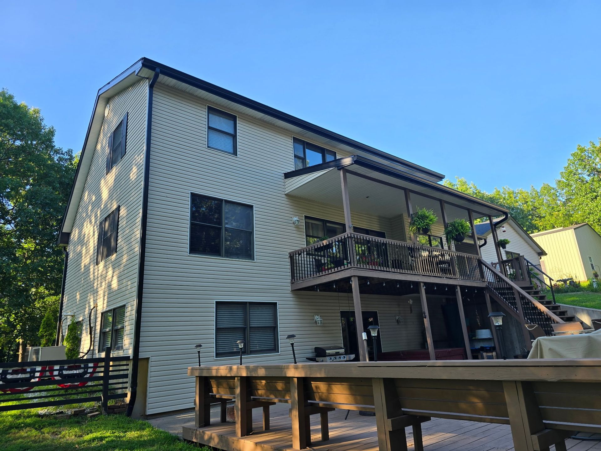 Two-story house with tan siding, black roof, and a wooden deck overlooking a yard and trees.