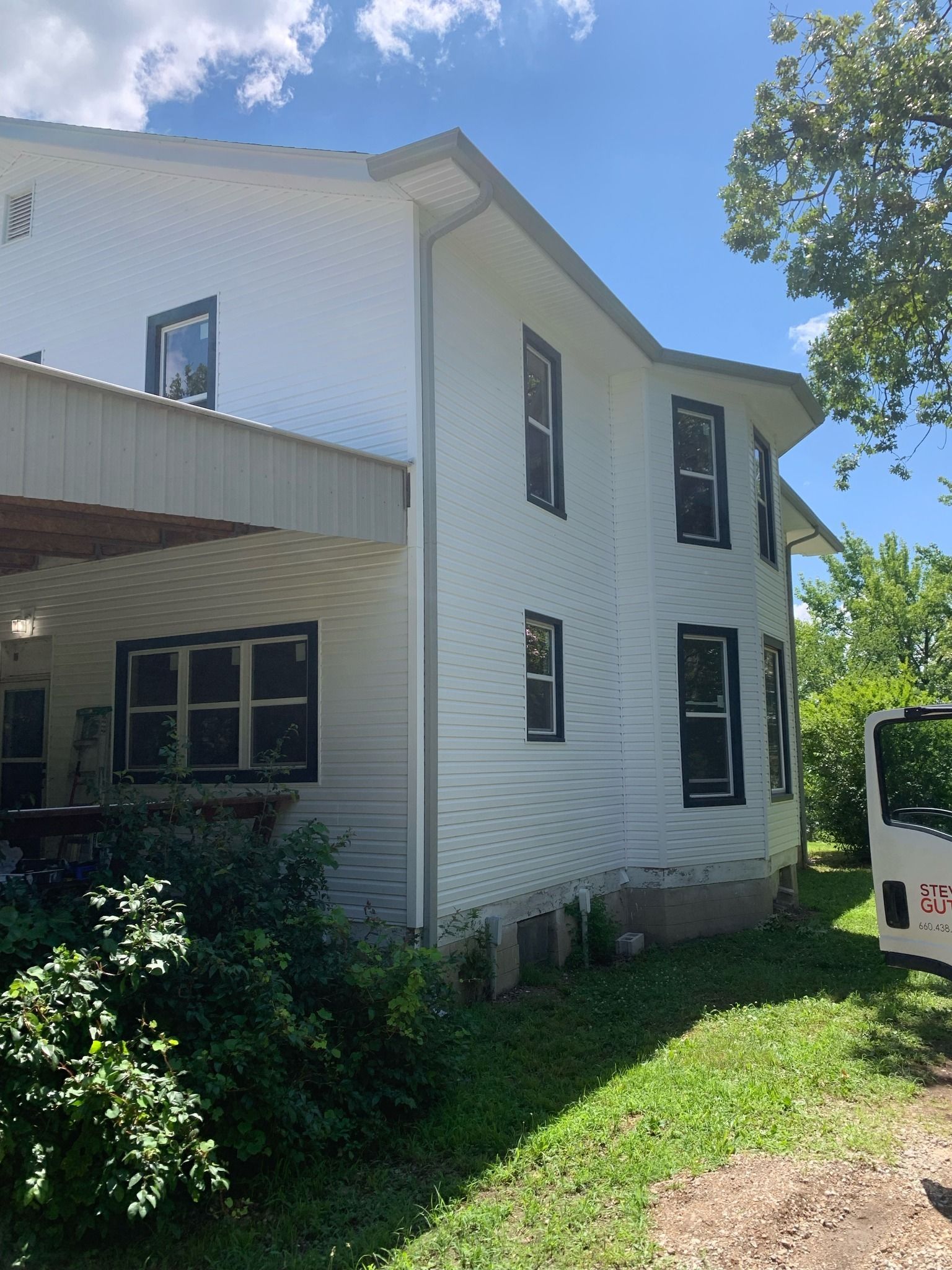 White two-story house with black-framed windows, surrounded by greenery, under a blue sky.