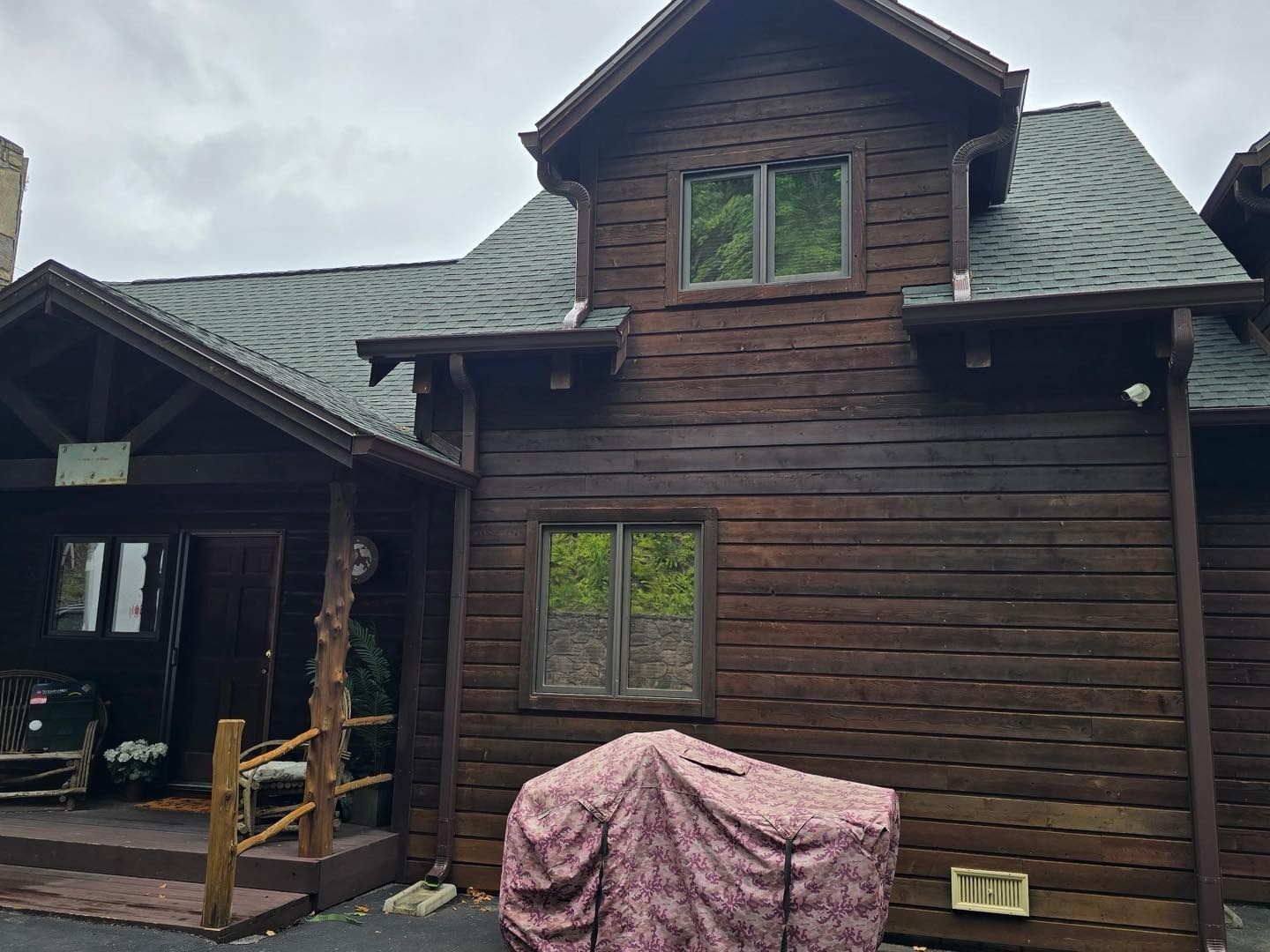 Wooden cabin with dark brown logs, porch, and two-story section with windows. Gray roof, overcast sky.