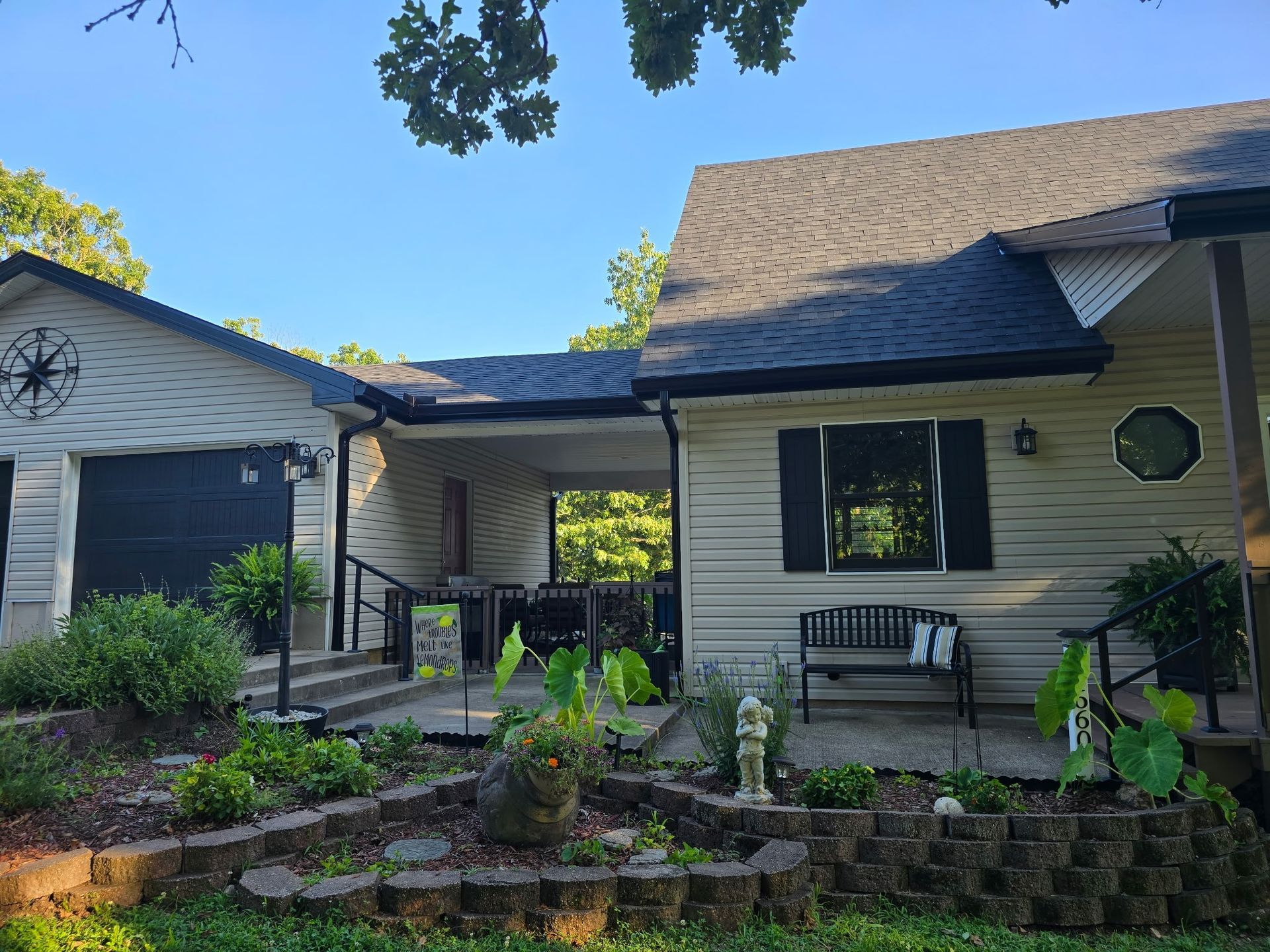 Beige house with black trim, a garage, porch, and a garden with plants.