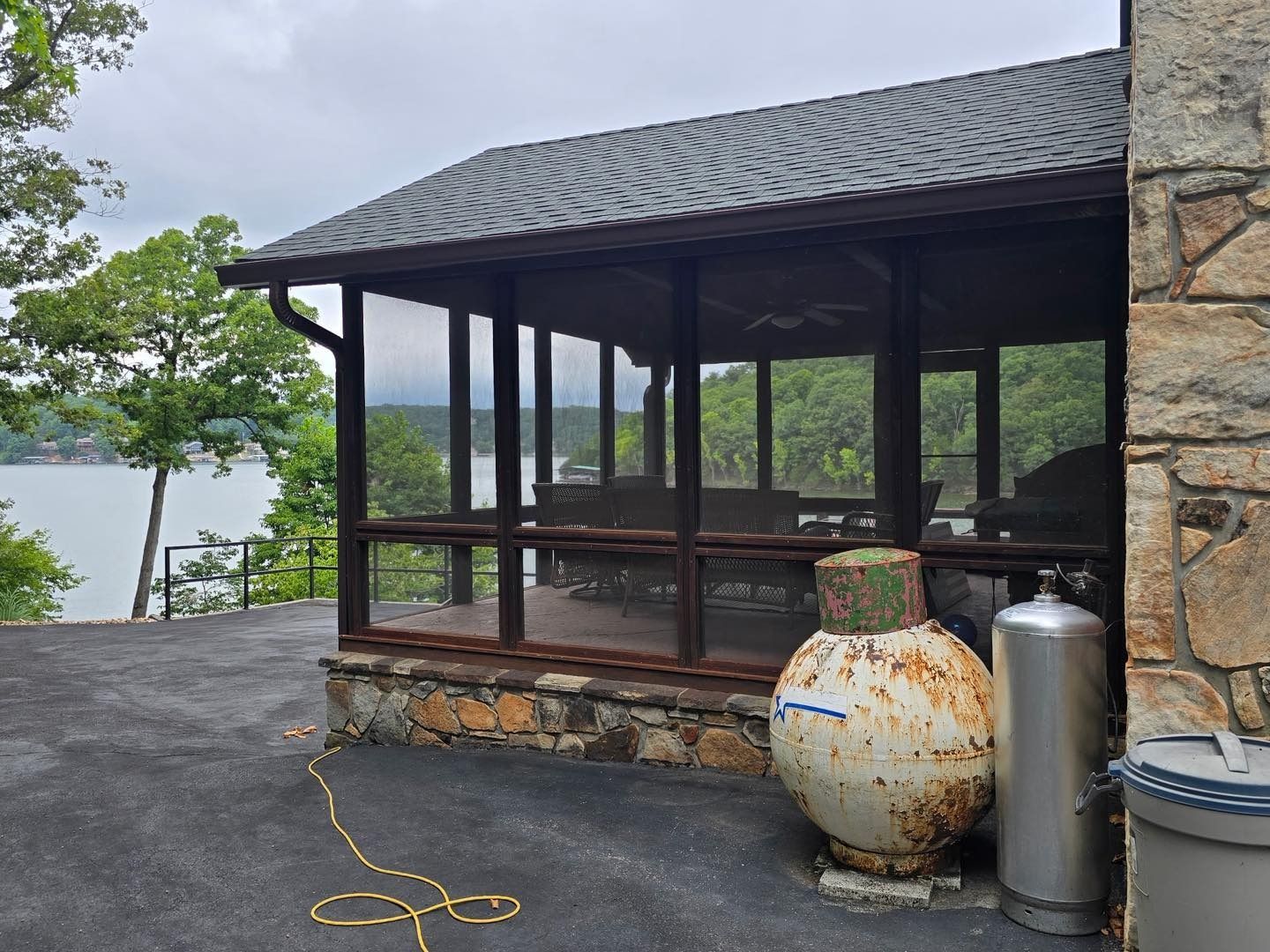 Screened-in porch with lake view, brown frame, stone base, dark roof, propane tanks in the foreground.