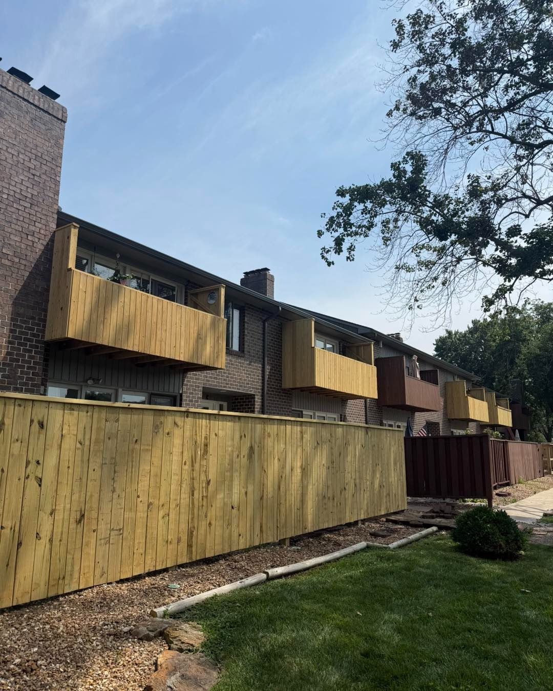 Row of townhouses with brown brick and tan balconies, behind a long tan wooden fence. Green grass and blue sky.