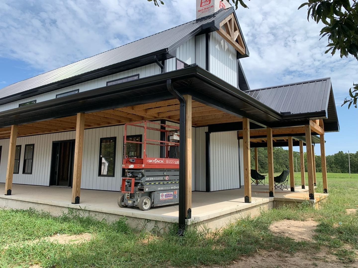 A modern farmhouse with a black metal roof, white siding, and a wrap-around porch; a lift sits on the porch.