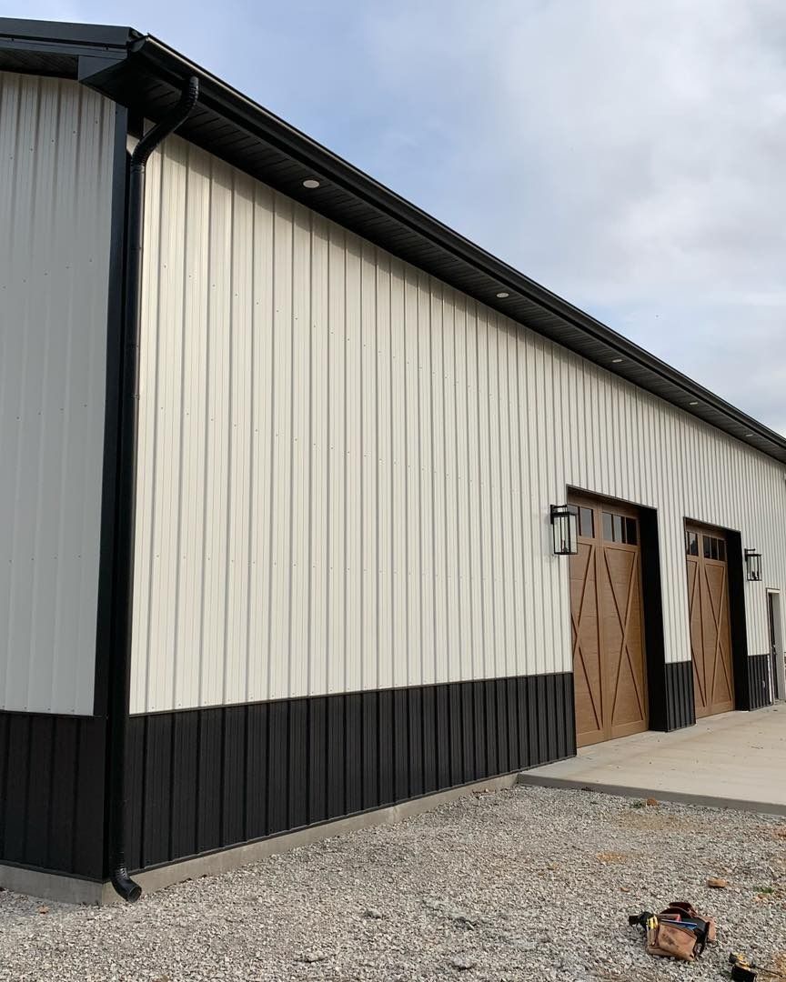 White and black metal building with brown doors. Black gutters and downspout. Gravel ground.