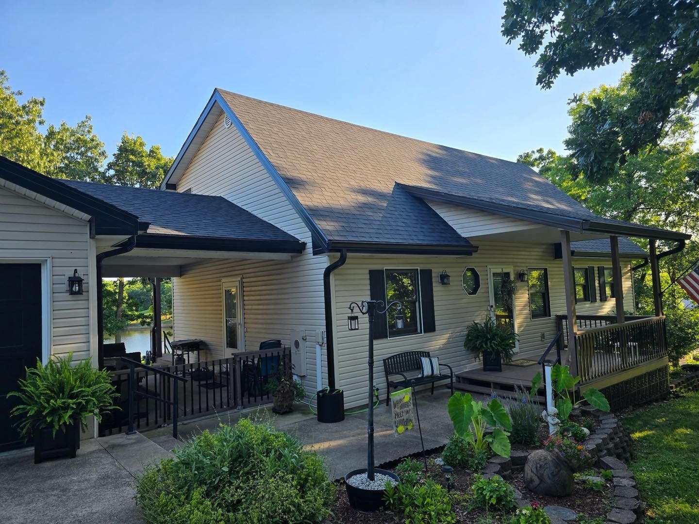 Tan house with a dark roof and porch, next to a waterfront with lush greenery.