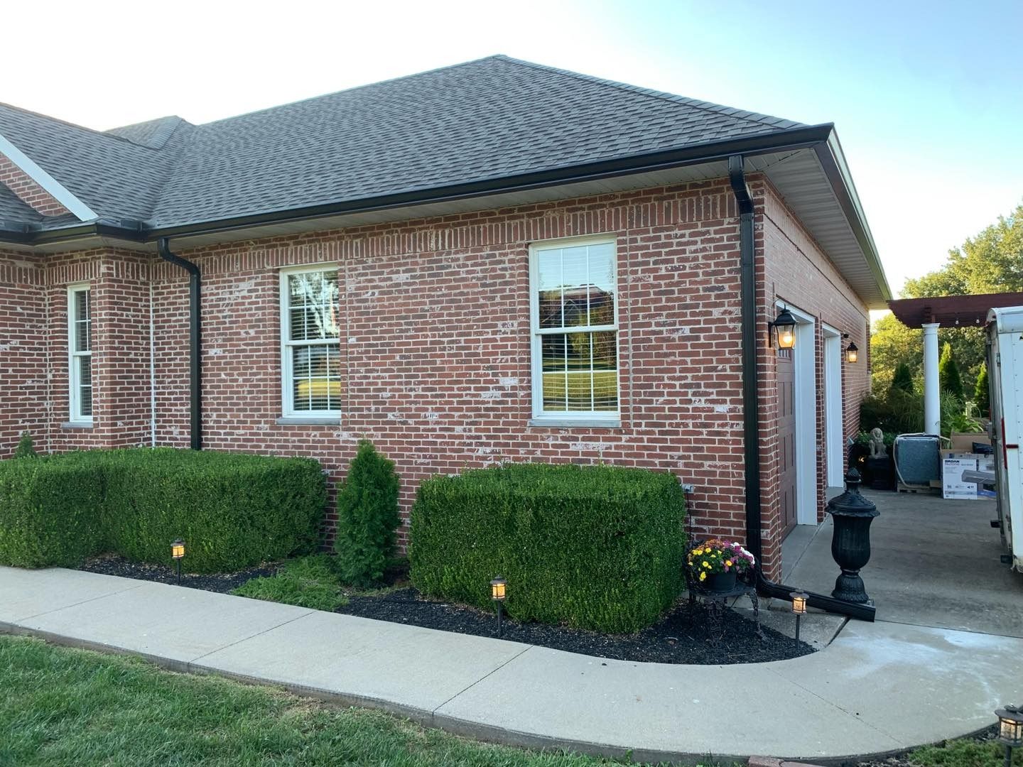 Brick house with green shrubbery, windows, and black gutters. Driveway visible.