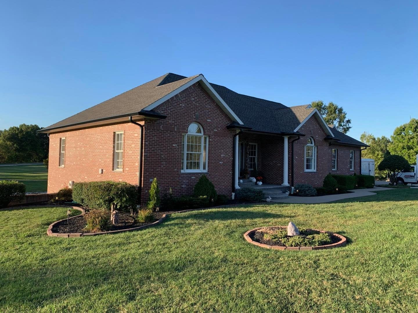 Brick house with dark roof and landscaping on a grassy lawn under a clear blue sky.