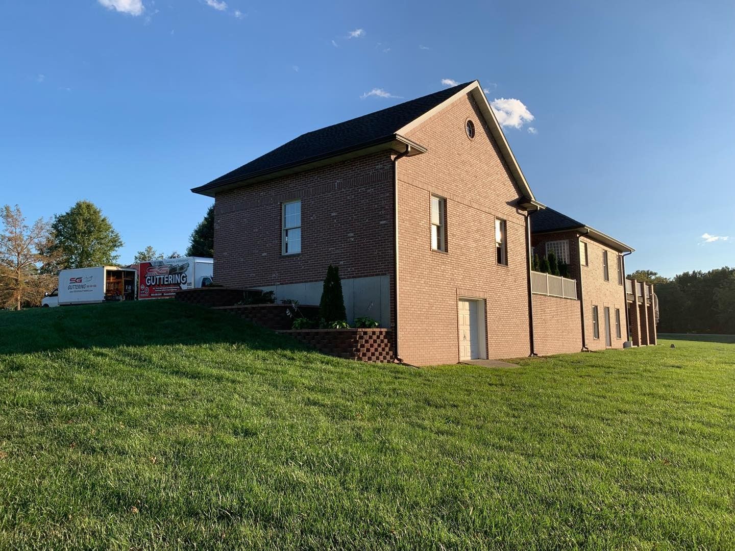 Brick house on a grassy hill; blue sky in the background. White moving containers on the left.