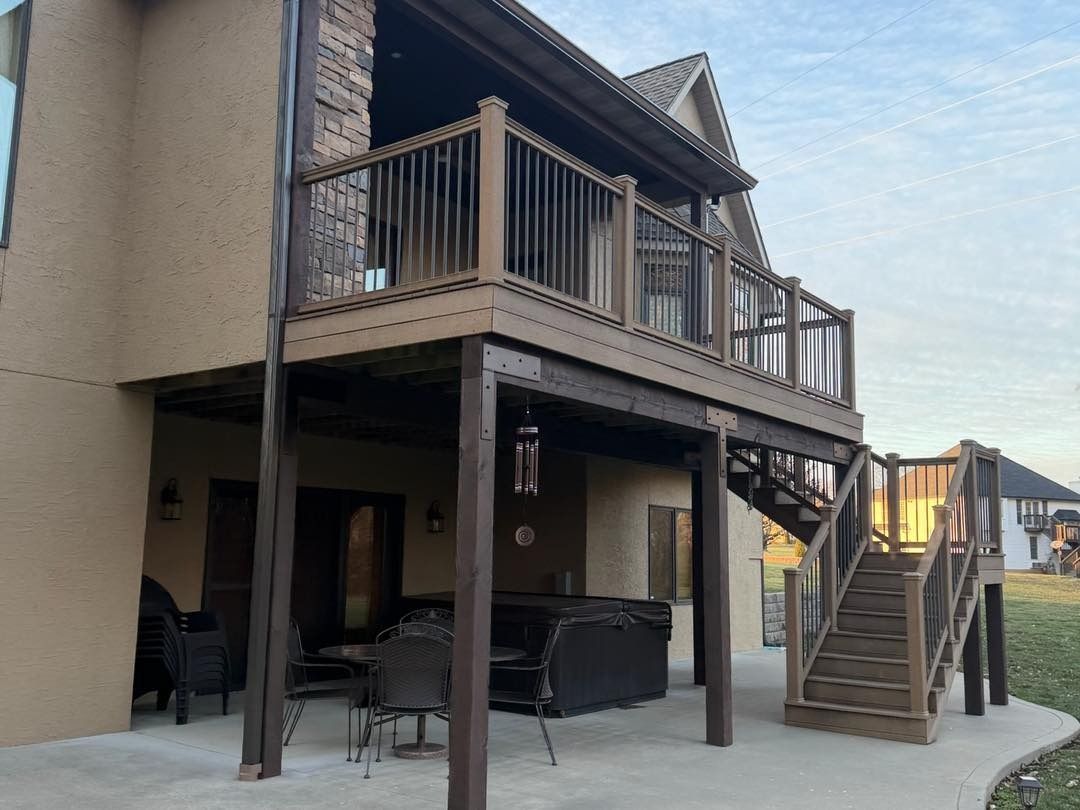 Two-story house with brown deck, stairs, and stone chimney. Tan exterior, concrete patio.