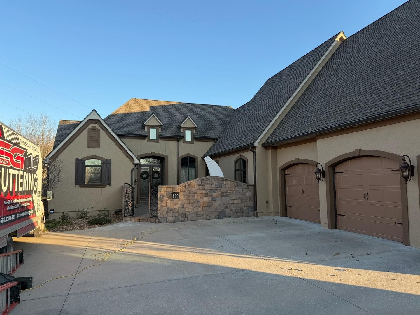 Tan stucco house with brown roof and garage doors, stone wall, and clear sky.