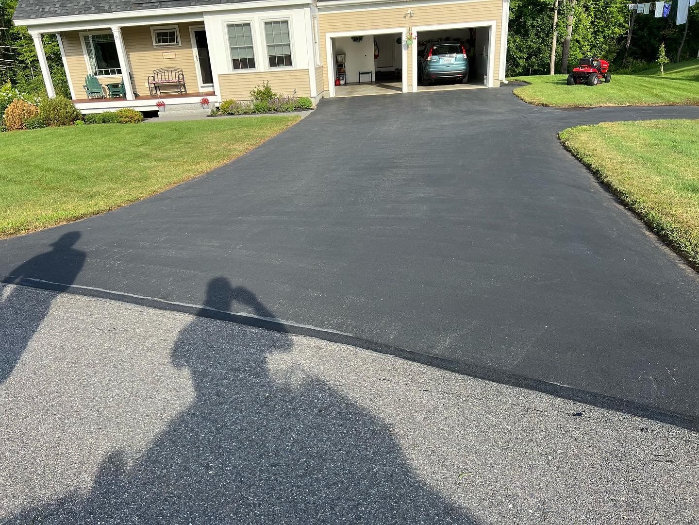 Newly sealed black asphalt driveway leading to a garage, connected to a gravel road and surrounded by green grass.