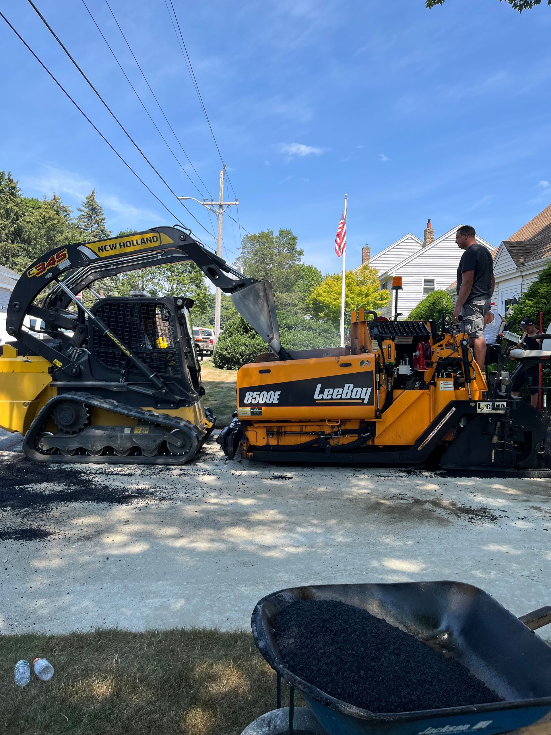 A skid steer loading asphalt into a paving machine on a sunny street, a person stands nearby.