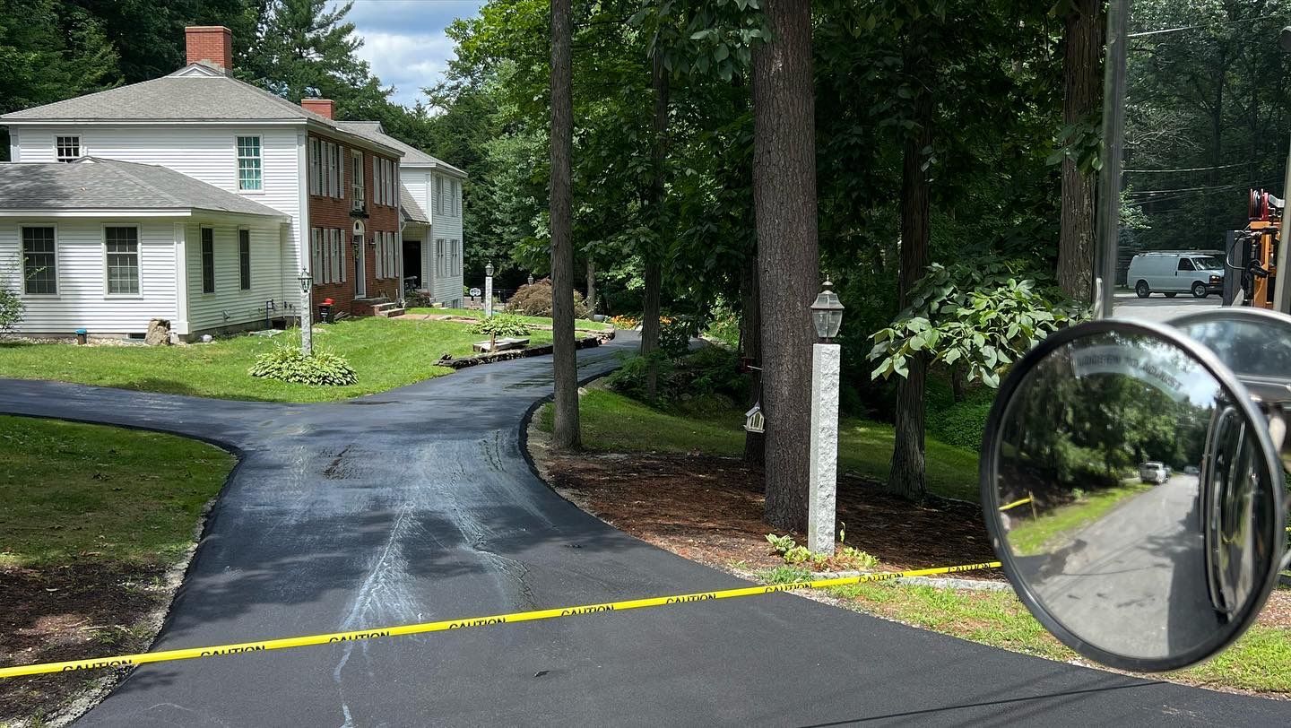 Newly paved asphalt driveway curves past a white and brown house. Trees line the right side.