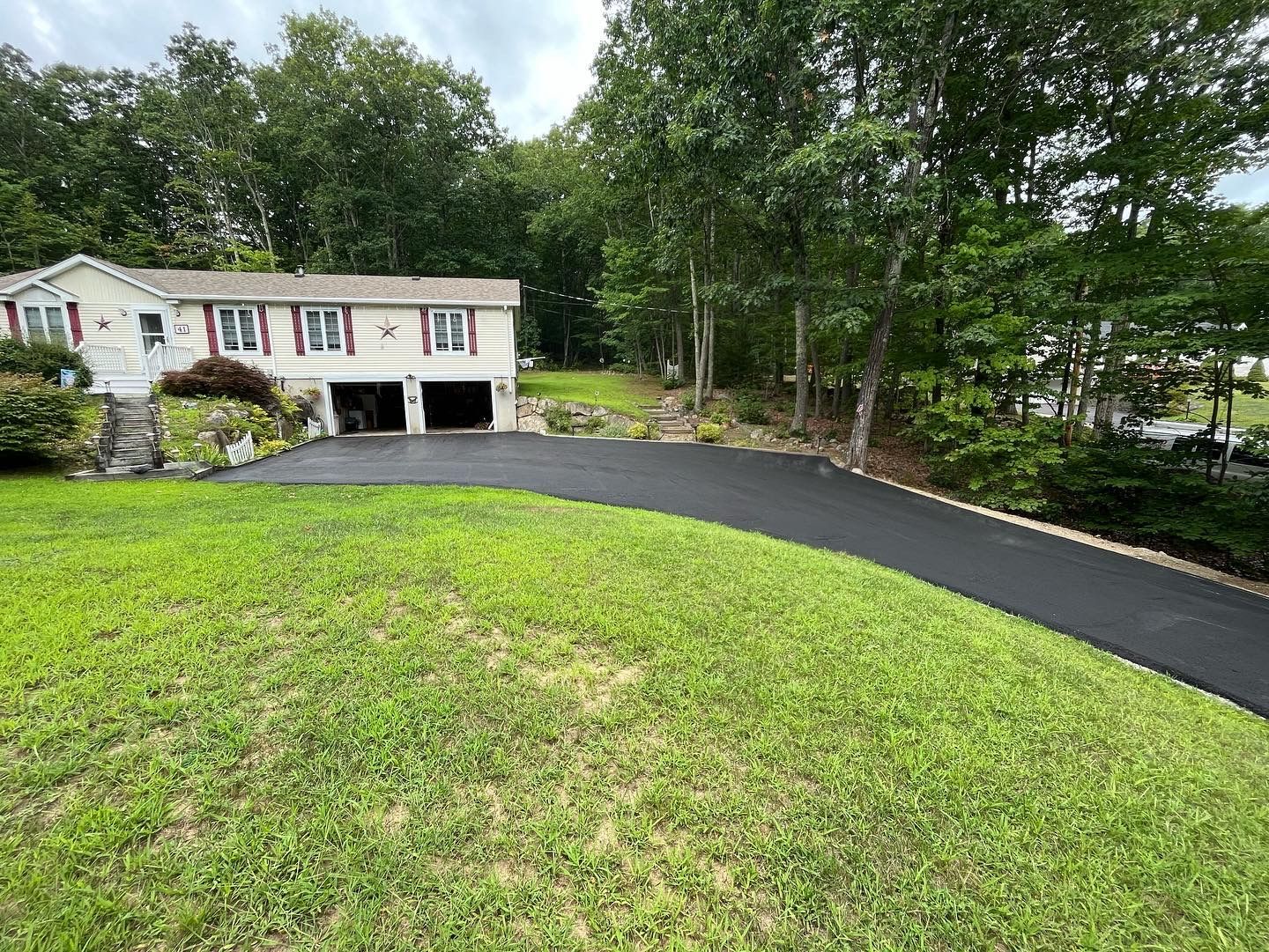 House with newly paved driveway and green lawn surrounded by trees.