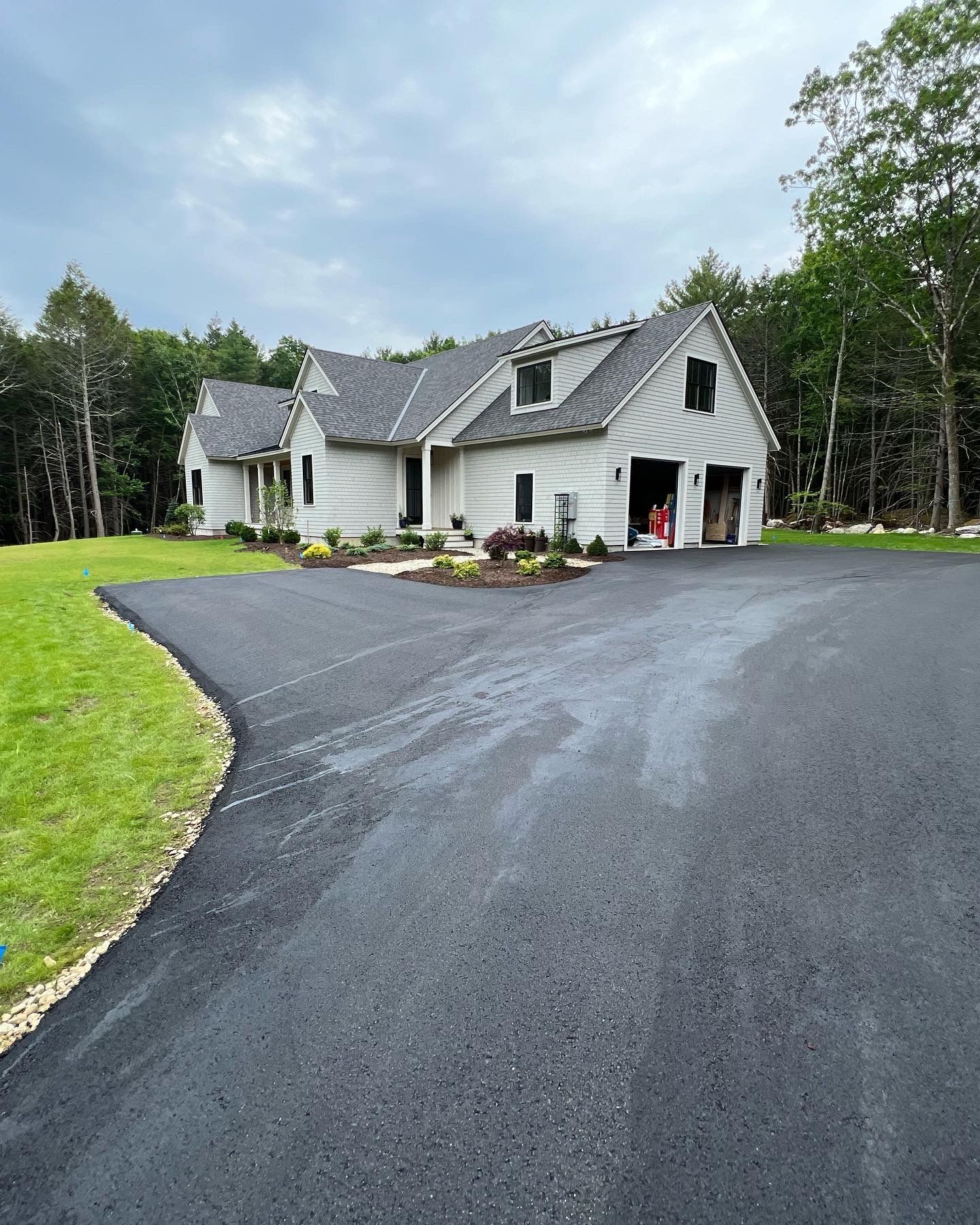 Light gray house with asphalt driveway and green lawn, cloudy sky, surrounded by trees.