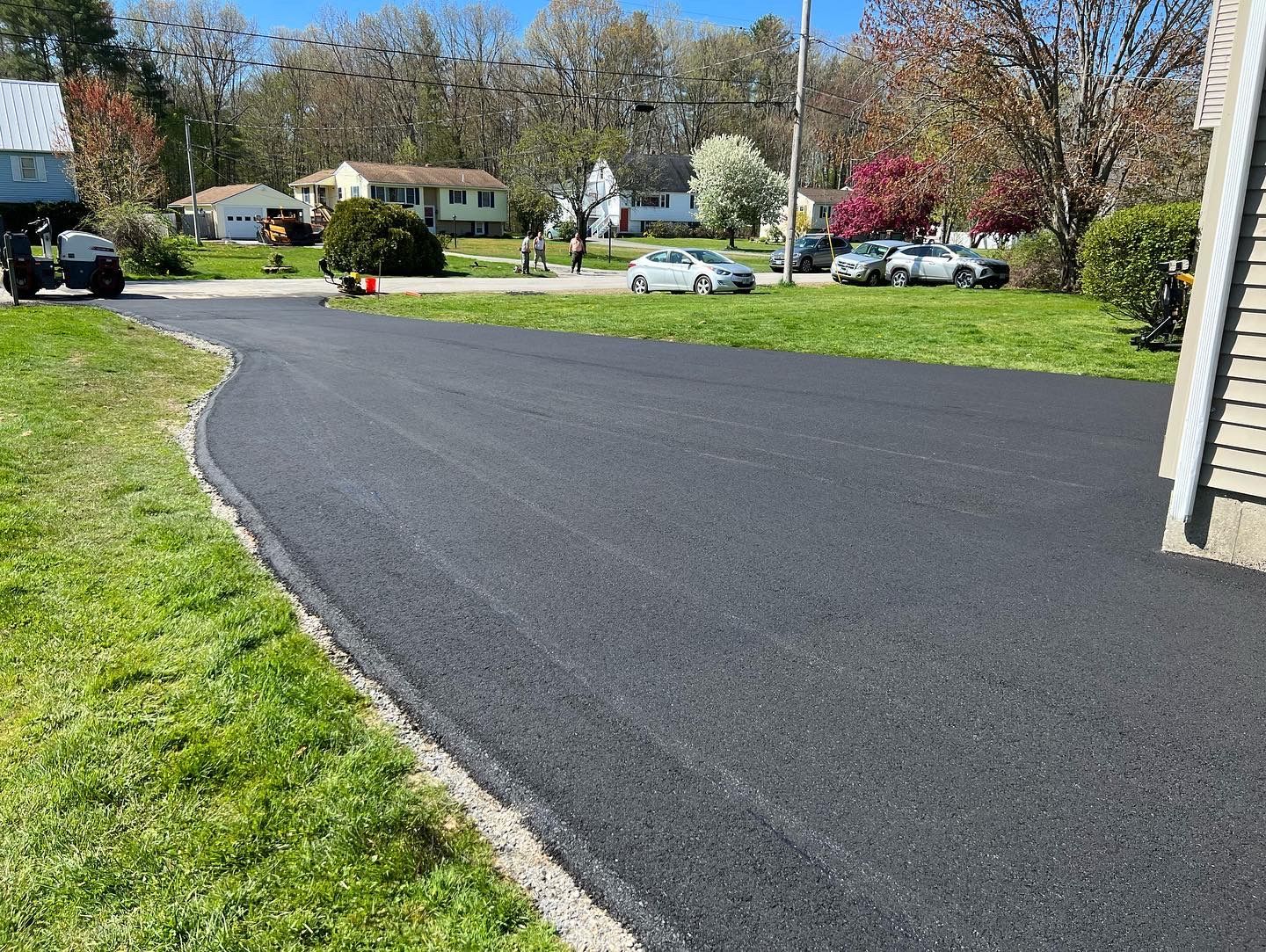 Newly paved asphalt driveway curves through a residential neighborhood with green grass and parked cars.