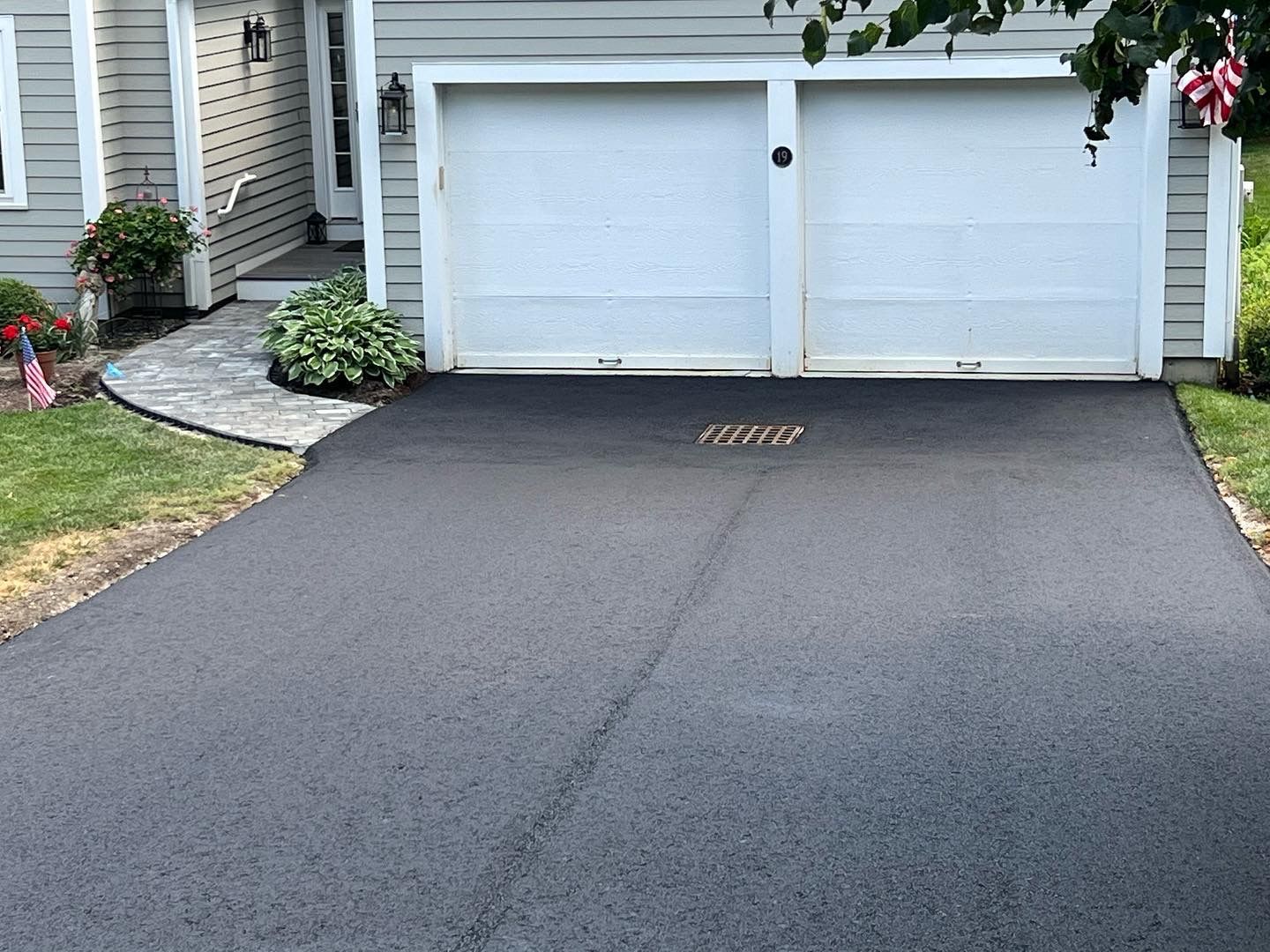 Black asphalt driveway leads to a two-car garage with white doors. Green lawn and a small garden are on the left.