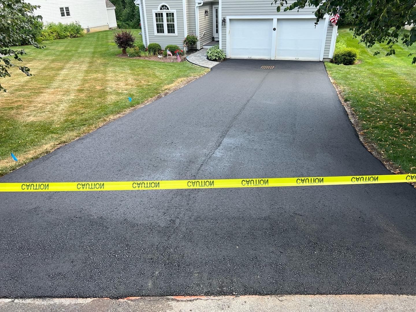 Freshly paved asphalt driveway with yellow caution tape, leading to a house with a two-car garage.