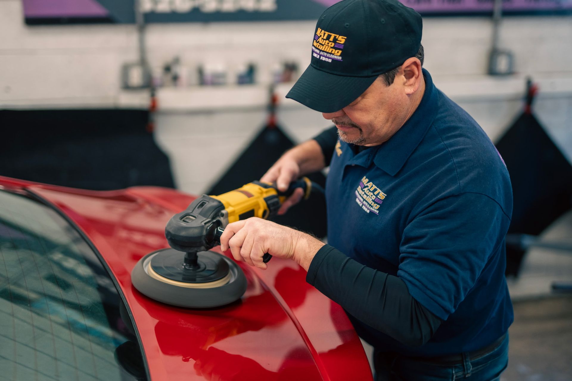 A worker in a navy uniform polishes the red hood of a car with a yellow electric buffer in a garage.