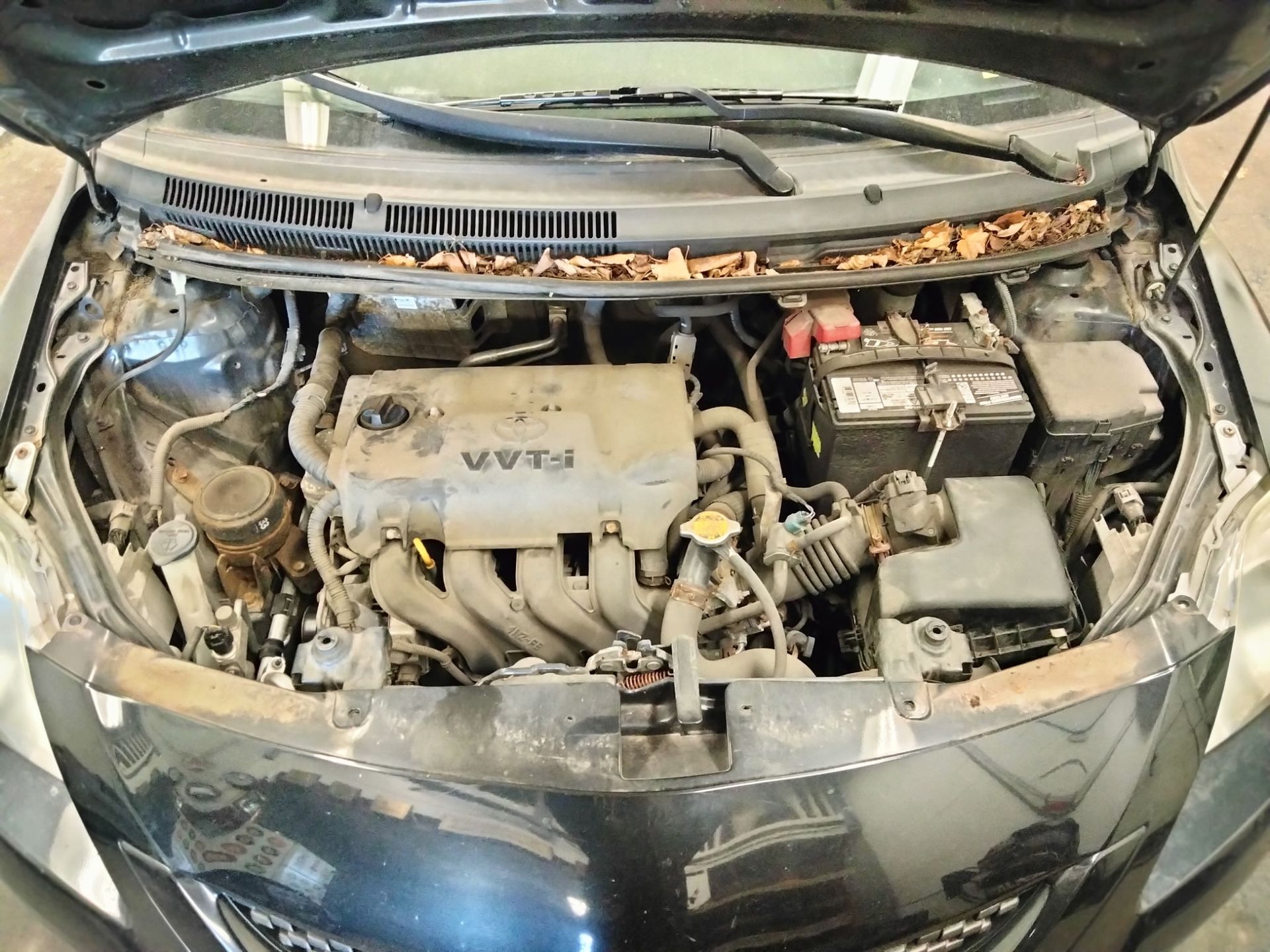 An open hood of a black car revealing a dusty engine bay with a VVT-i engine, car battery, and debris near the windshield.