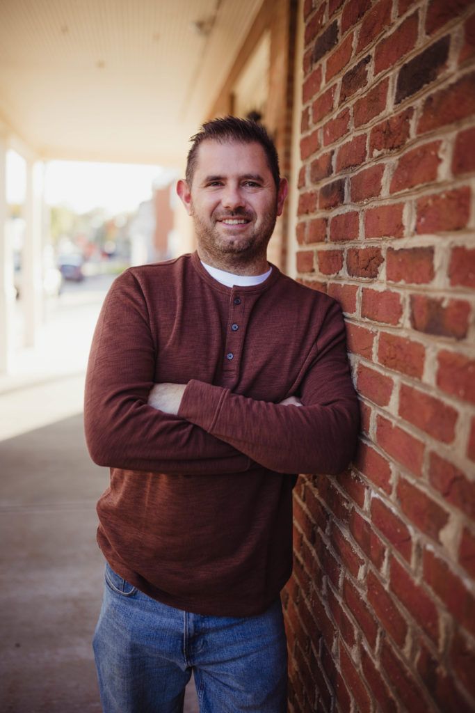 Man with arms crossed leaning against a brick wall, wearing a maroon shirt and jeans, smiling.