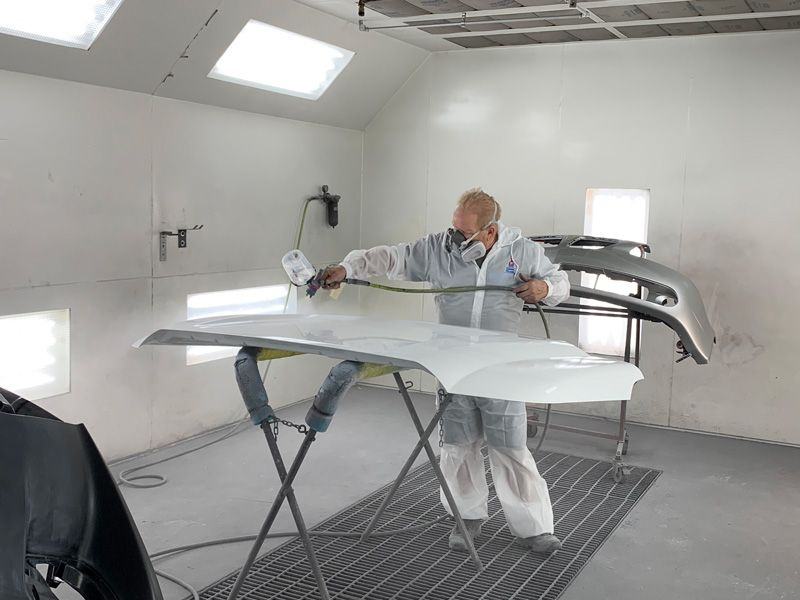 Person in protective suit sprays white paint on car hood in a paint booth.