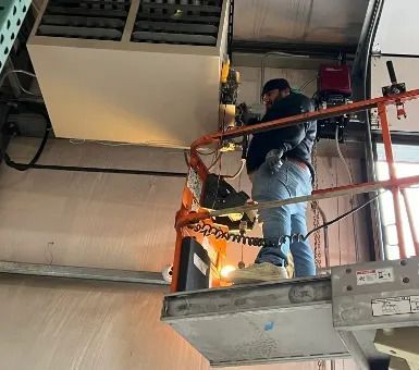 Worker on a lift repairing overhead equipment inside a building, with sparks flying.